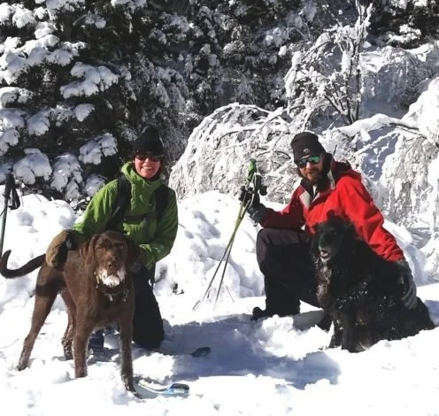 Two people in winter gear with two dogs on a snowy mountain, mountains and snow-covered trees in the background.