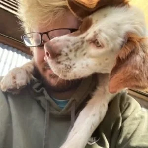 Person with glasses hugging a white and brown spotted dog.
