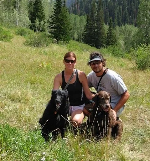 A man and woman kneeling on grass in a forested area, each with a dog, with trees and mountains in the background.
