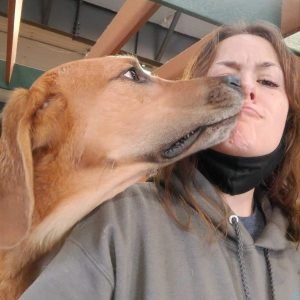 Deedra making a kissing face while a dog licks her face.