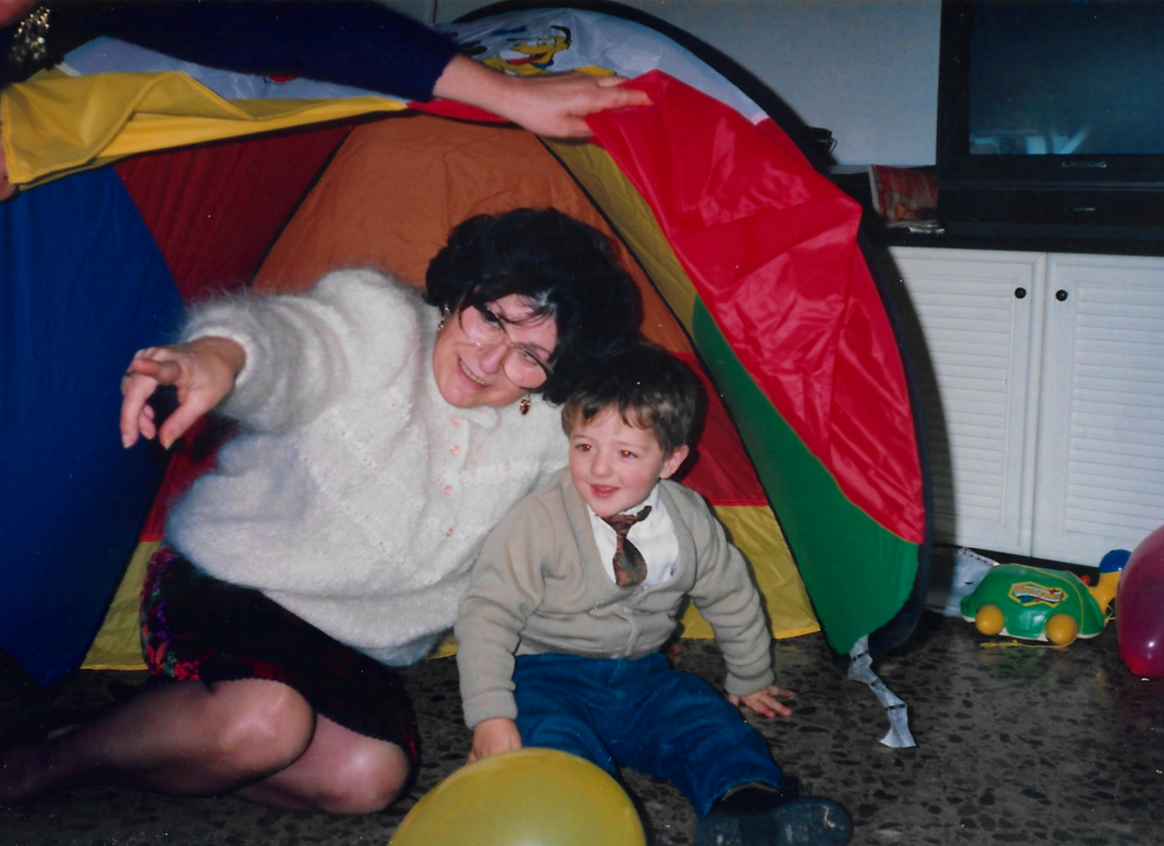 Una mujer y un niño pequeños jugando y sonriendo dentro de una tienda de campaña colorida en una sala de estar, rodeados de globos y juguetes.