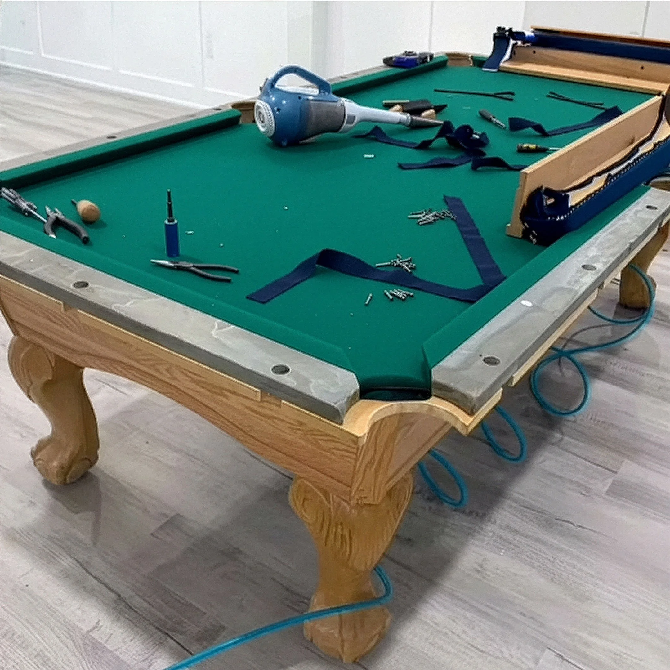A green felt pool table with various tools and objects scattered on its surface, including a caulking gun, a screwdriver, pliers, screws, a lanyard, and some elastic straps. The table has a wooden base with ornate legs, and some blue cords or hoses are hanging underneath.