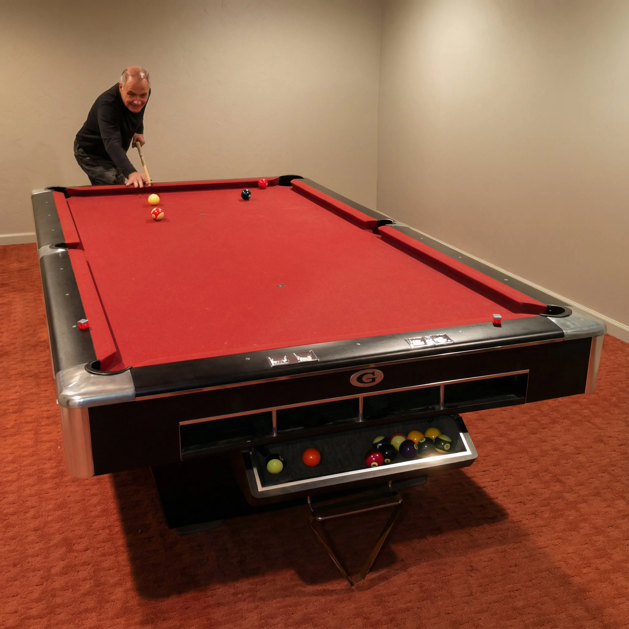 A man smiling while leaning over a red pool table in a room with beige walls and reddish carpeting, preparing to take a shot.