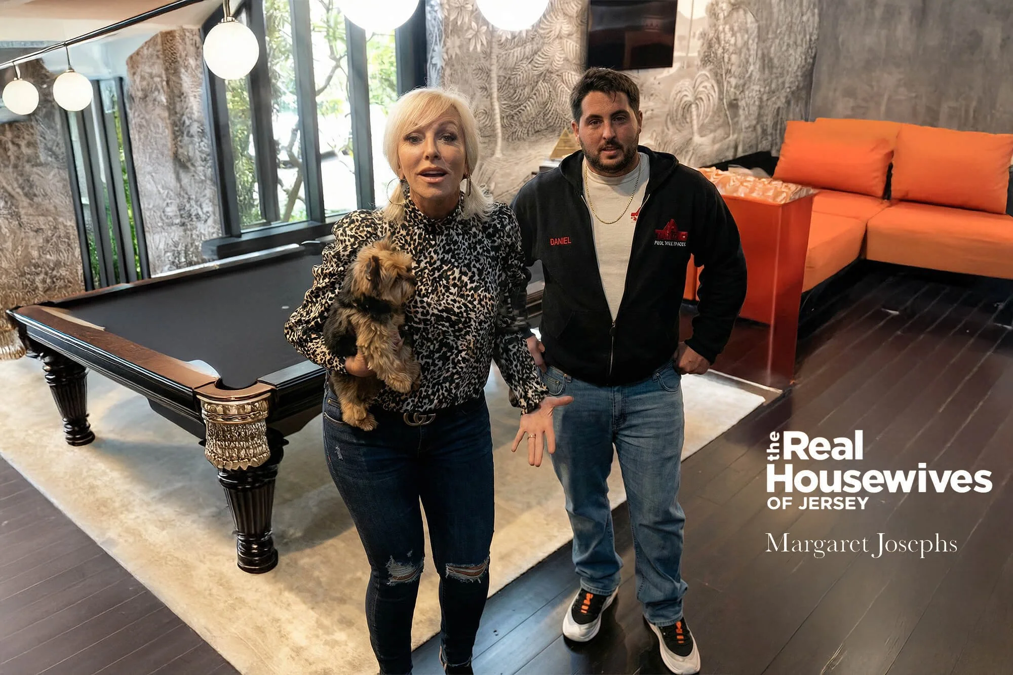 Two women and a man standing in a living room with a pool table, large windows, and modern furniture. The woman on the left is holding a small dog and is wearing a leopard print top, while the man on the right is dressed casually in a hoodie. The ima