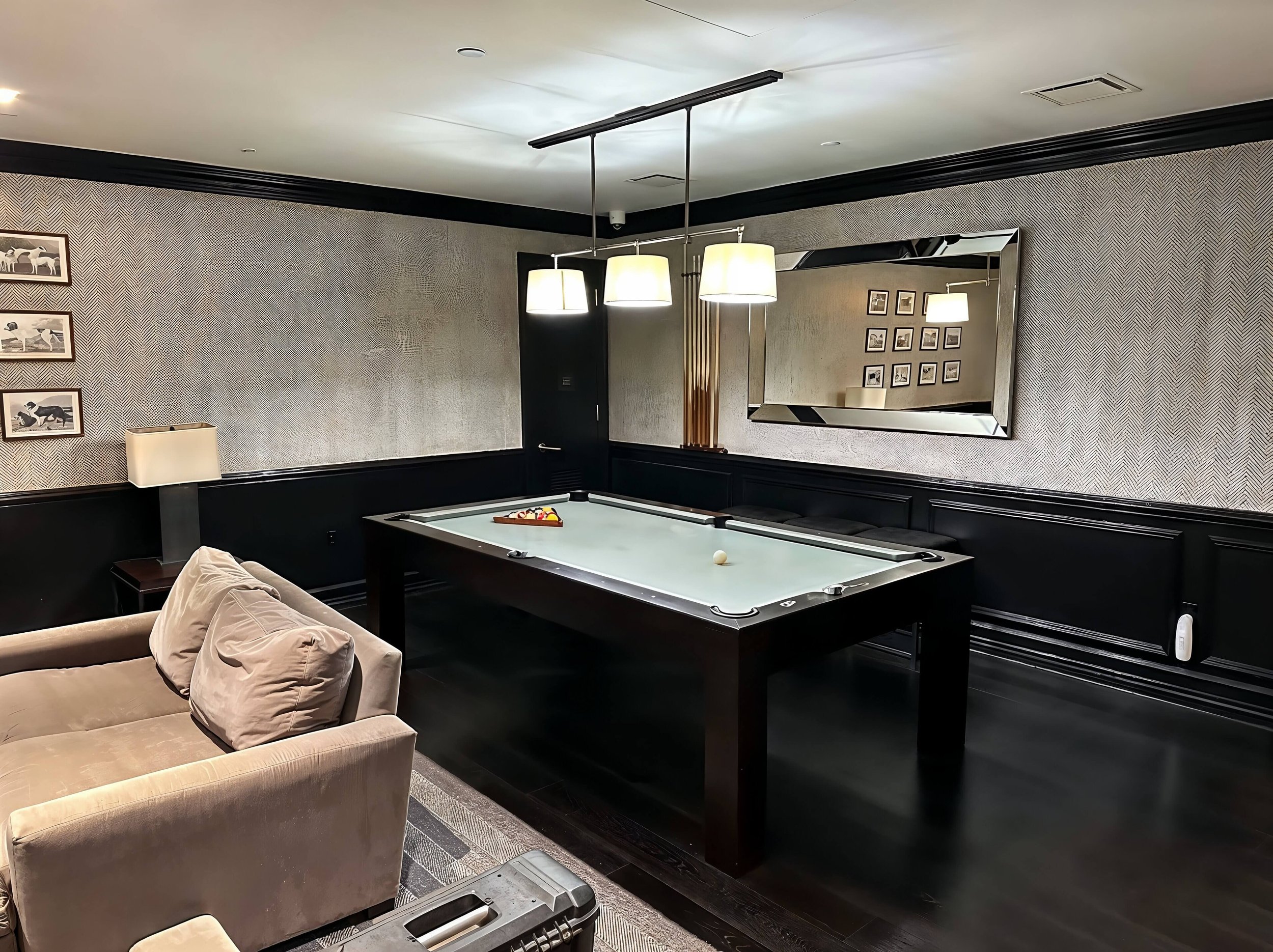 A room with a pool table, beige sofa, wall mirror, framed photos, and two table lamps.