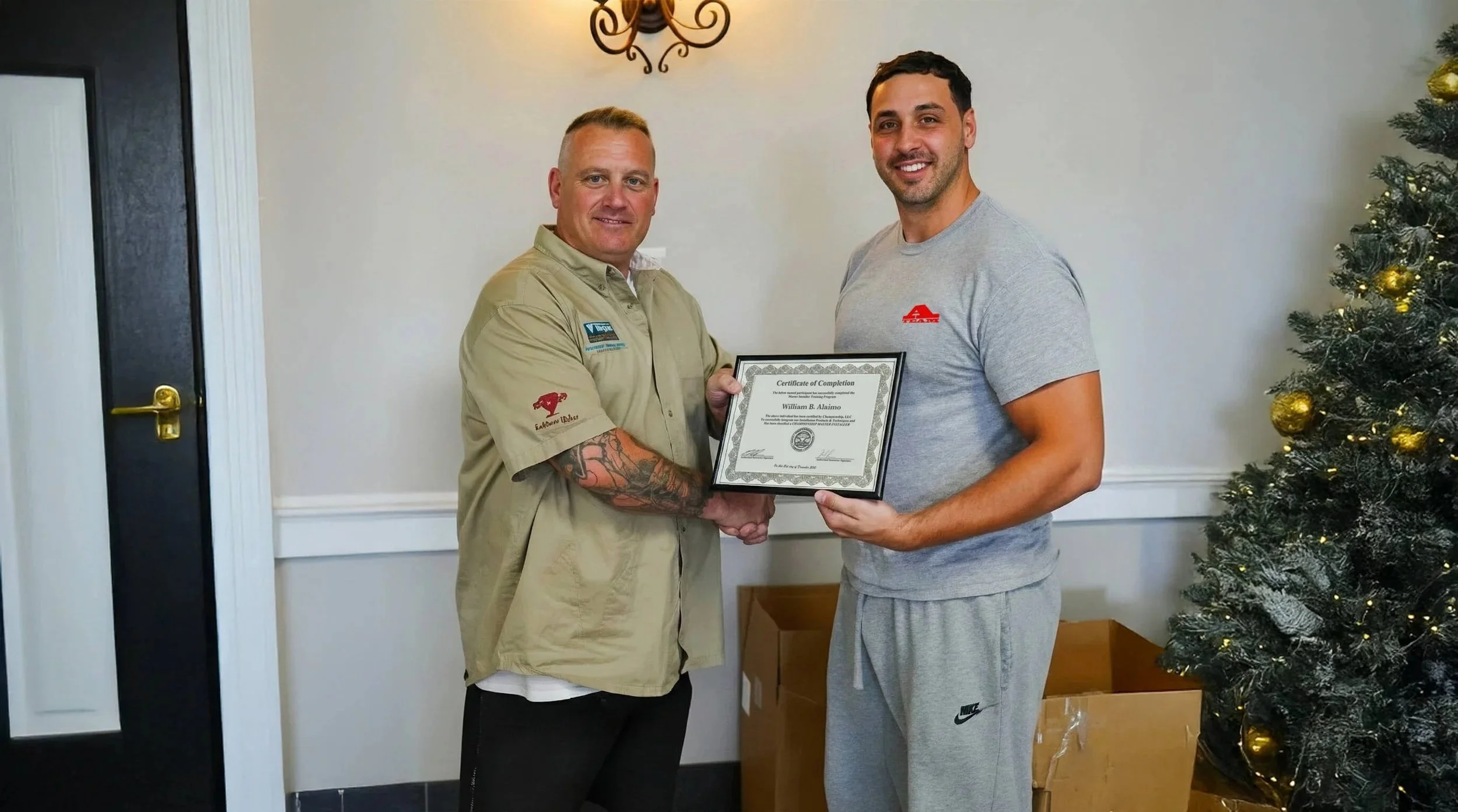 Two men standing indoors, shaking hands, with one holding a framed certificate. The man on the left has tattoos, is wearing a tan shirt with patches, and is smiling. The man on the right is in a gray shirt and sweatpants, also smiling. There is a dec