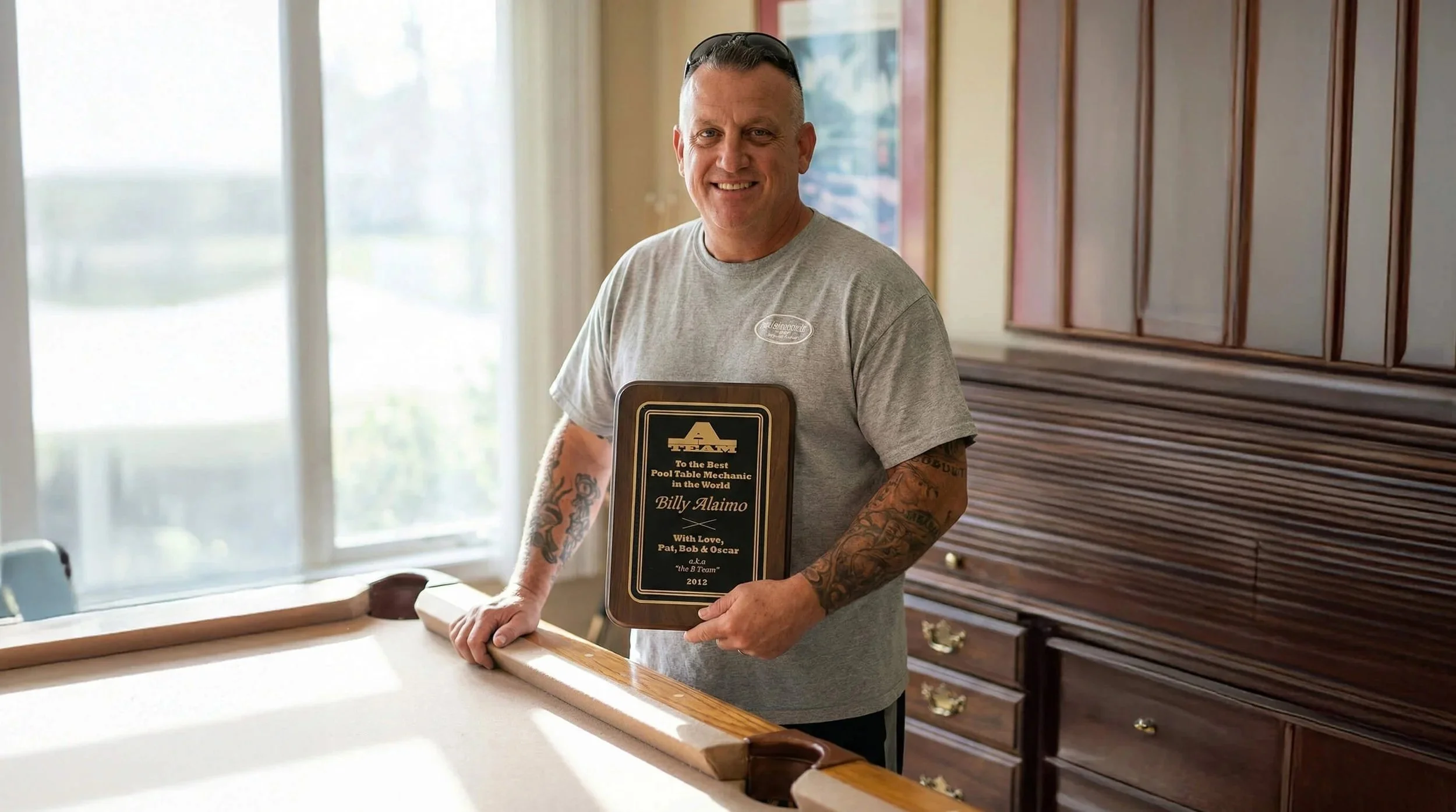 A man with short dark hair, tattoos on both arms, wearing a gray t-shirt, stands beside a pool table holding a plaque, smiling at the camera inside a room with wooden furniture and large windows.