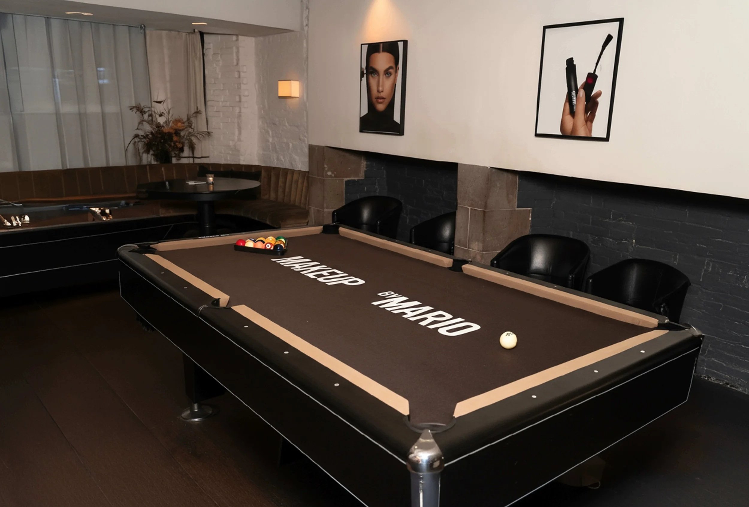 Pool table with a set of billiard balls, cue ball, and stacked cues in a dimly lit lounge with black chairs, framed advertisements on the wall, and a corner seating area with a table and decorative plant. Make Up Mario Pool Table Design by  A-Team.