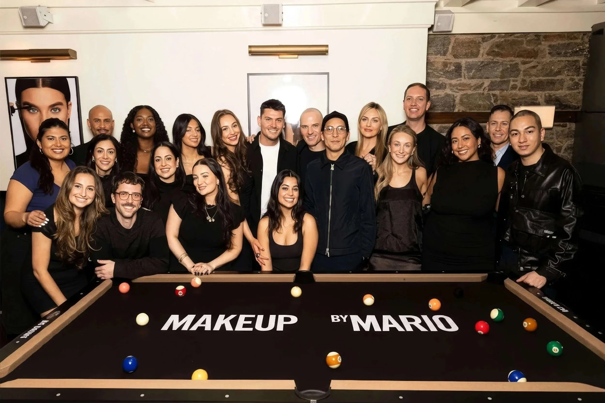 Group of people surrounding a pool table with the text 'MAKEUP BY MARIO' written on it, posing for a photo indoors.