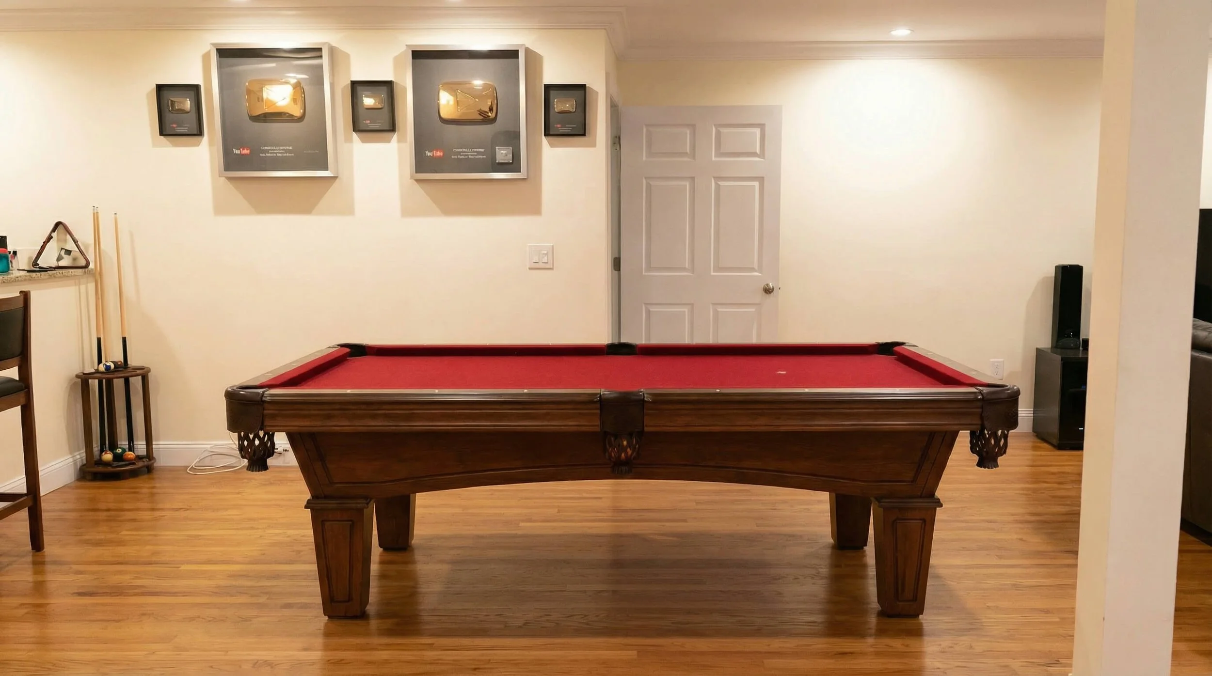 A wooden pool table with a red felt surface in a basement game room with hardwood floors. The wall behind has three YouTube award plaques, two in black frames and one large silver in the center, with a door on the right. To the left, a small rack wit