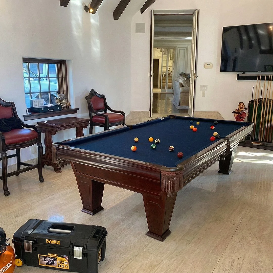 A room with a wooden pool table with pool balls on it, two red and dark wood chairs, a window with decorative items, and a TV mounted on the wall. There is an open door leading to another room, and a toolbox on the floor.