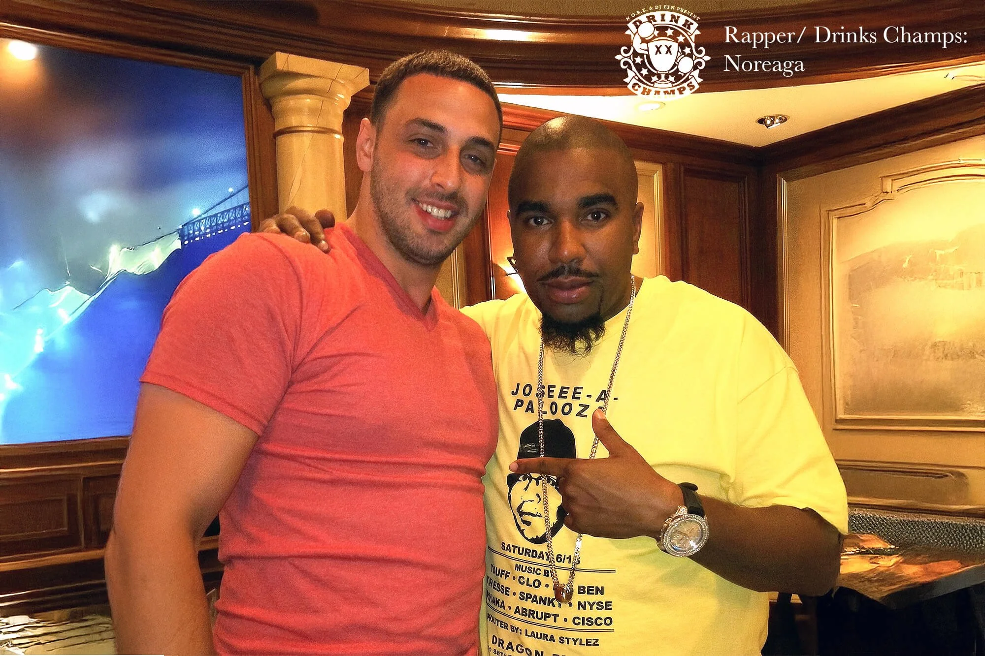 Two men posing together indoors; one in a red shirt with arm around the other in a yellow T-shirt pointing to him. Background shows wooden wall paneling and a large television screen. Drink Champs, Rapper Noreaga, N.O.R.E