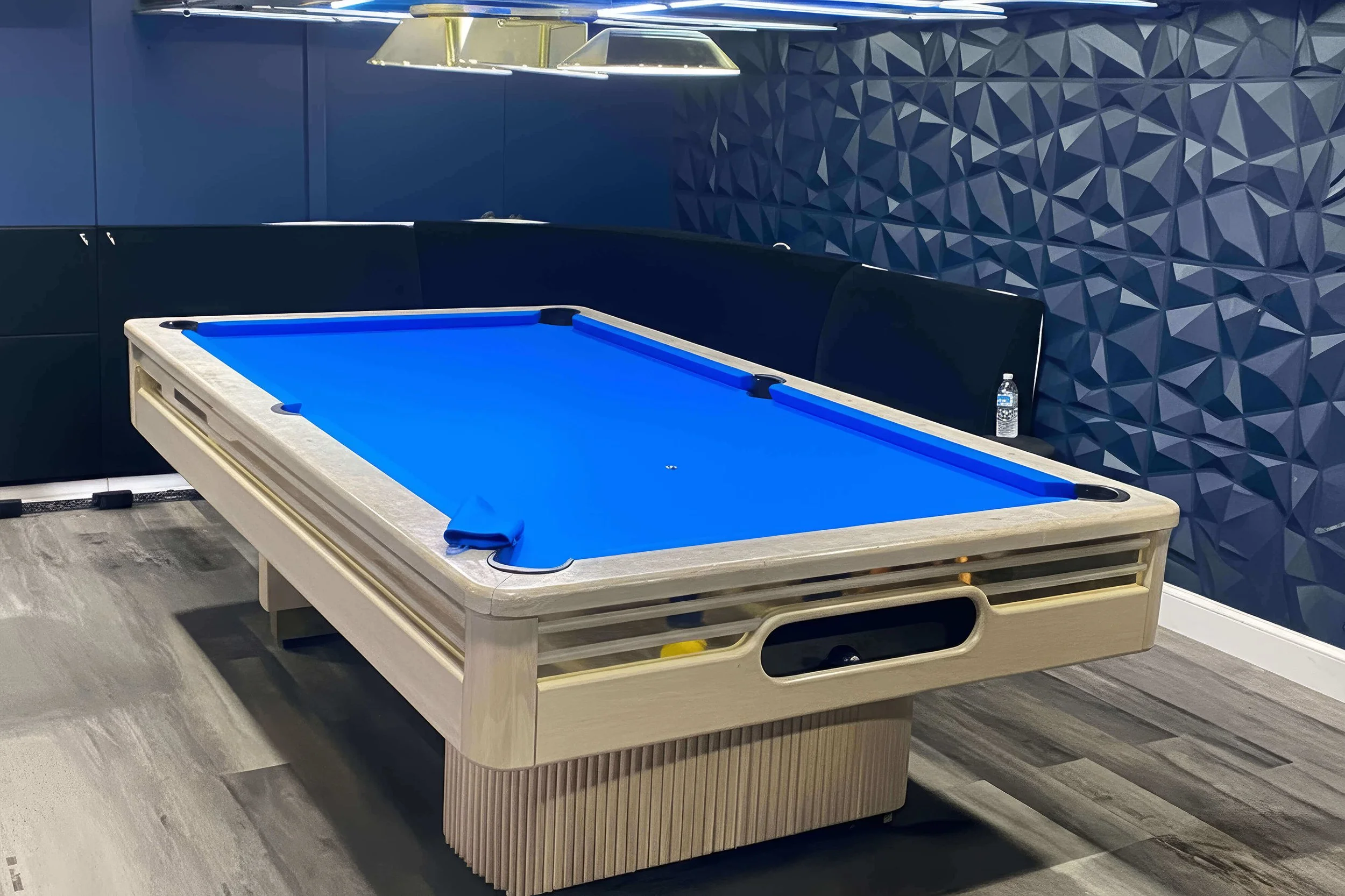 Modern billiard room with a pool table featuring a blue felt surface, black seating, a water bottle on the side, and a textured blue geometric wall.