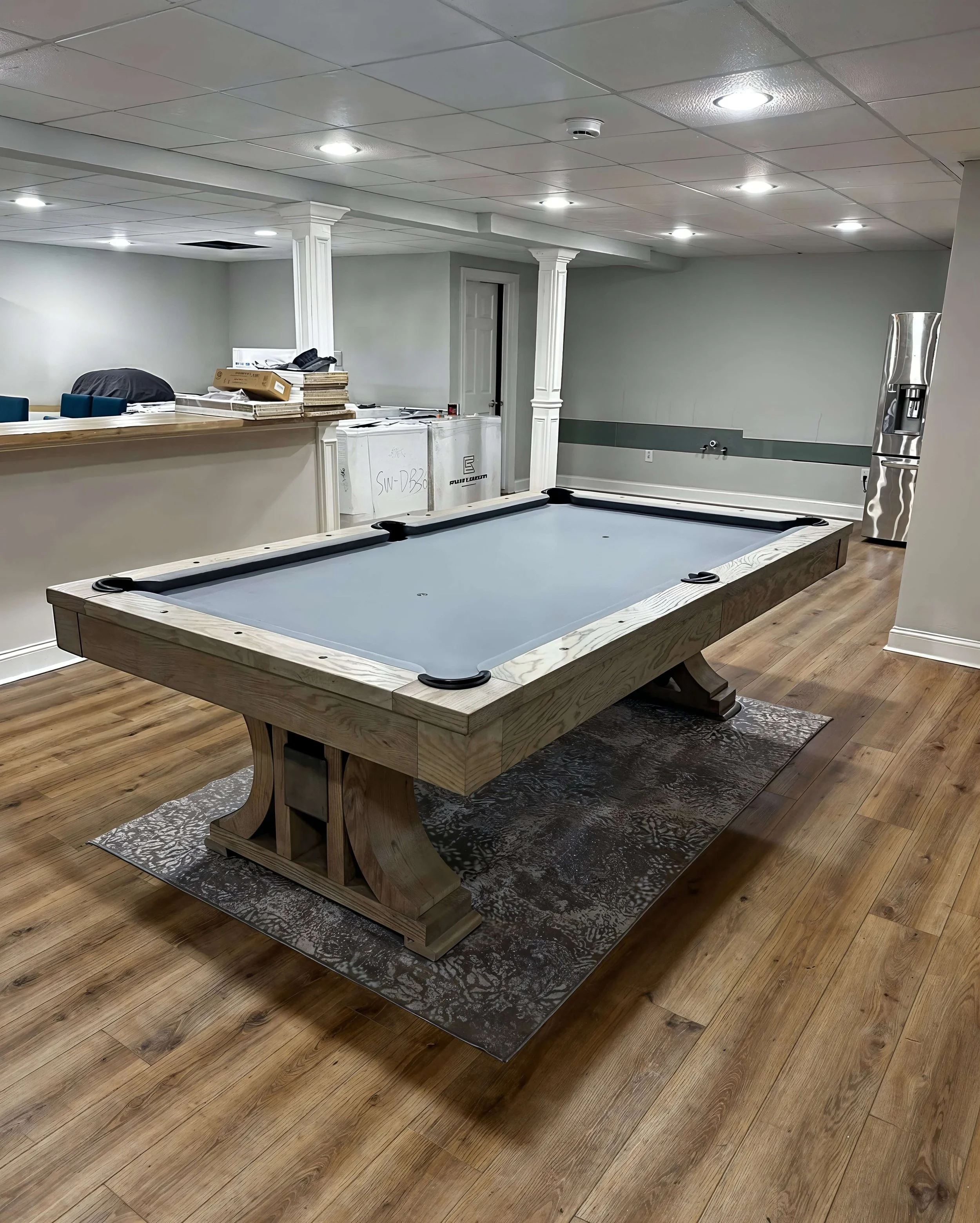 A home pool table with a light-colored wooden frame and a gray felt surface in a basement with wood flooring, white walls, and a water dispenser in the background.