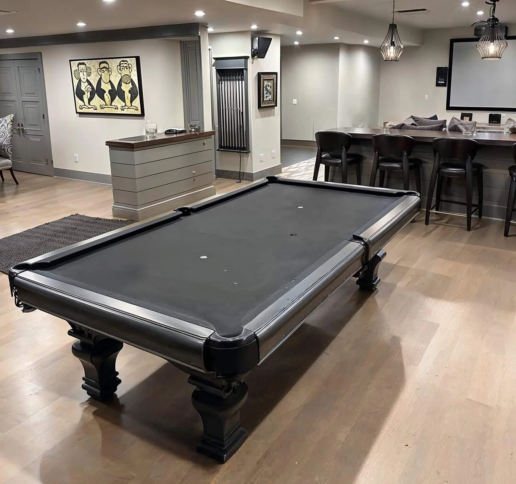 A modern indoor space with a pool table, bar counter with black chairs, wall art, and a large window or mounted screen in the background, illuminated by ceiling lights and pendant fixtures.