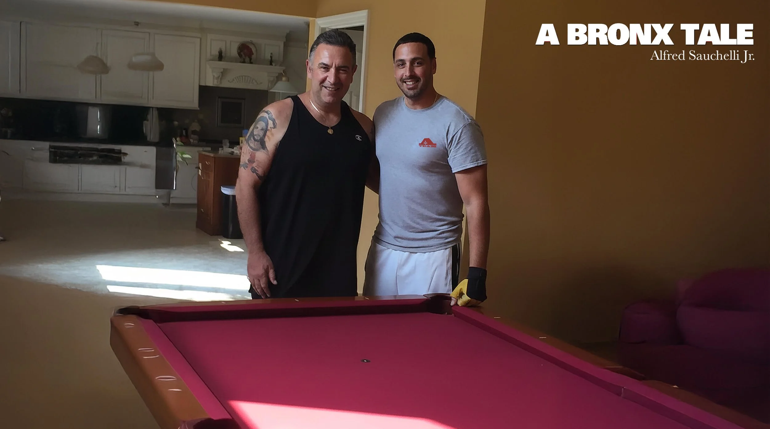 Two men standing together next to a pink pool table in a living room, smiling, with a kitchen visible in the background. Text on the image reads 'A BRONX TALE' and 'Alfred Sauchelli Jr.'