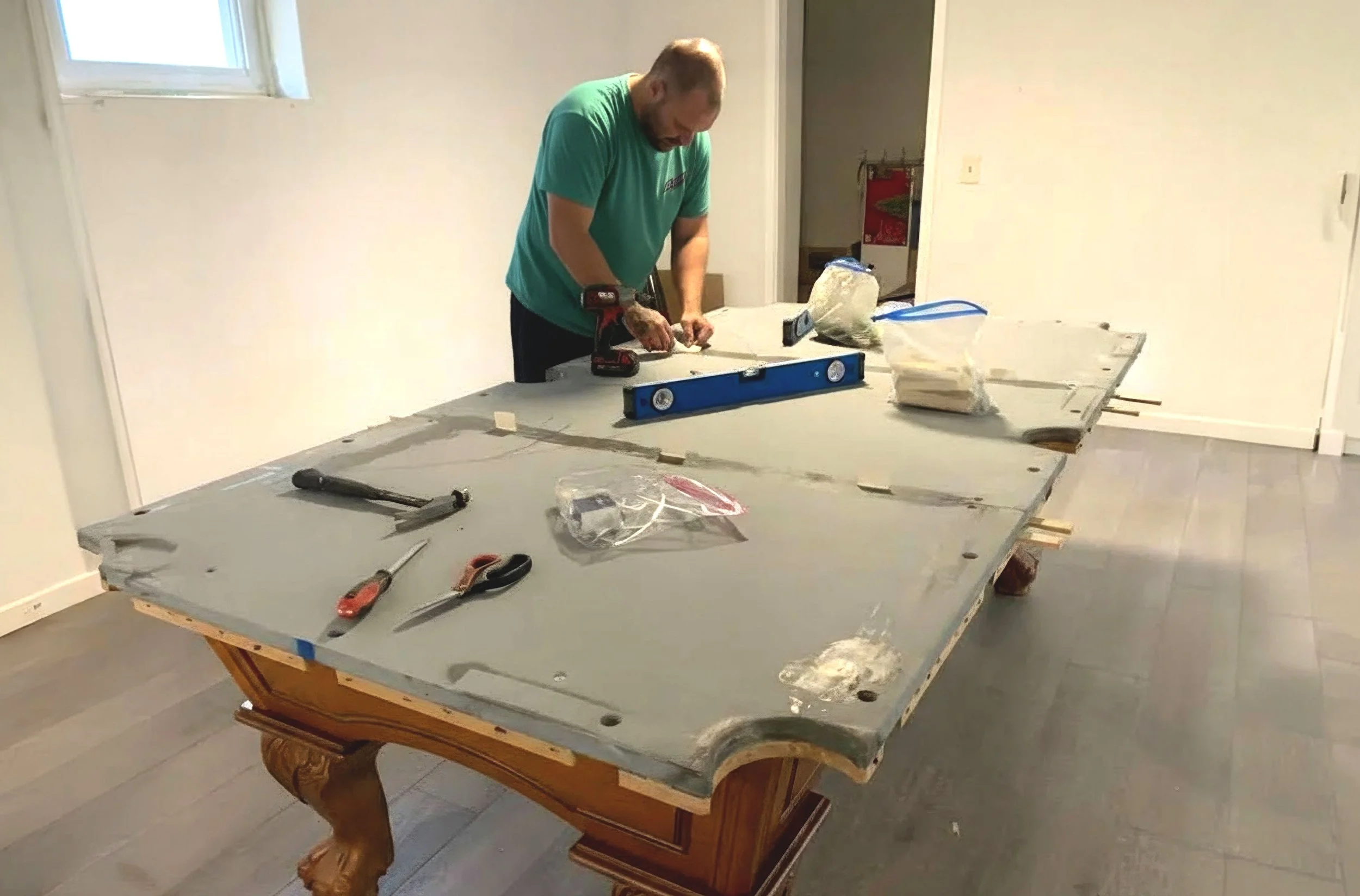 A man working on a pool table in an indoor room with light-colored walls and wooden flooring, using a level and tools on the table.