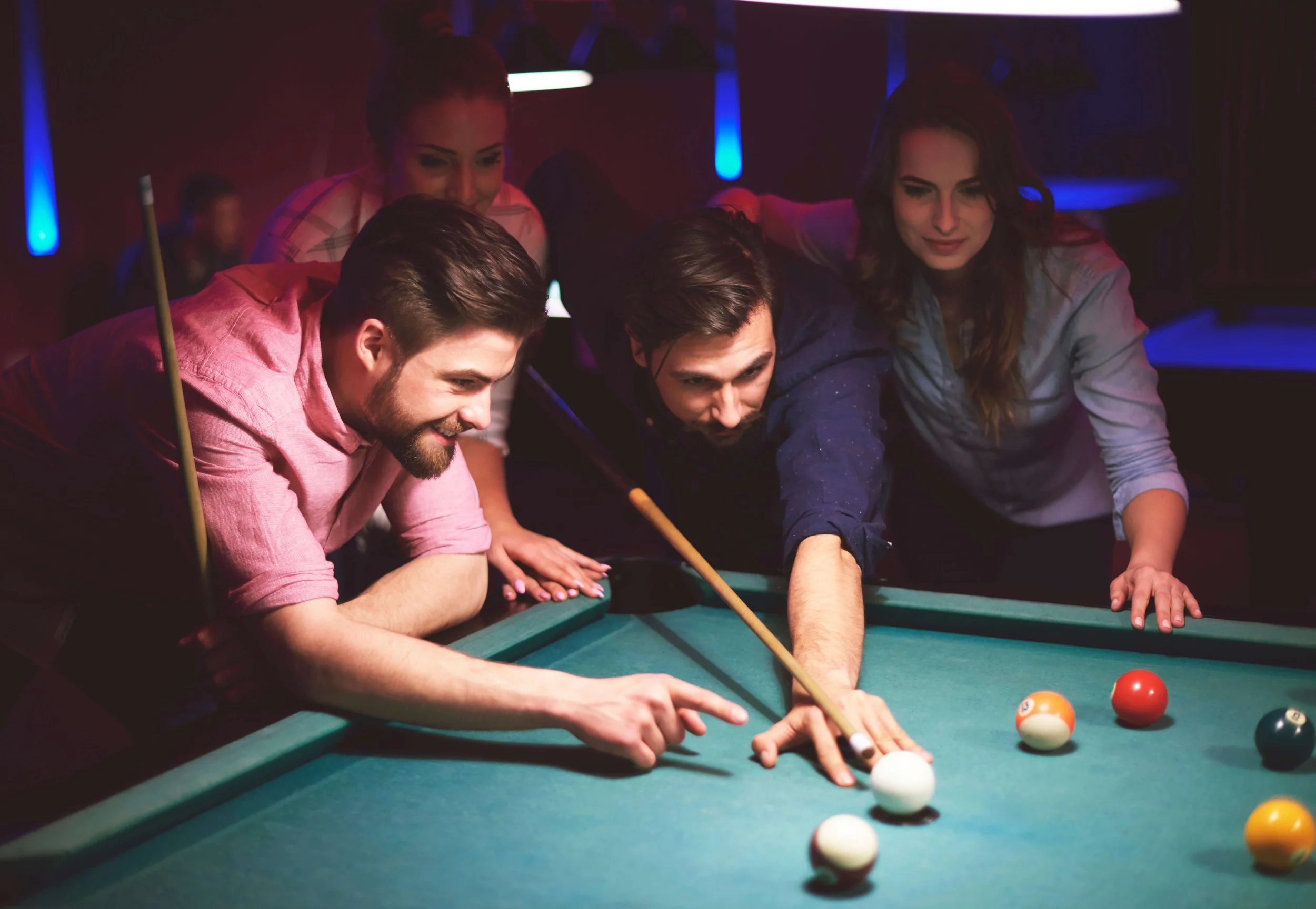Four friends playing pool at a dimly lit bar, focused on the game as they line up a shot