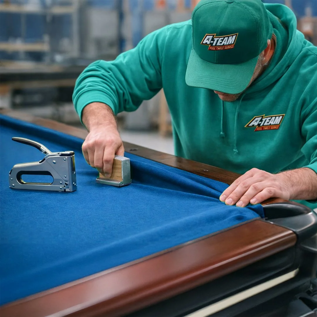 A man in a teal hoodie and cap with an A-TEAM logo installs blue felt on a pool table using a staple gun and a small tool for smoothing the fabric.