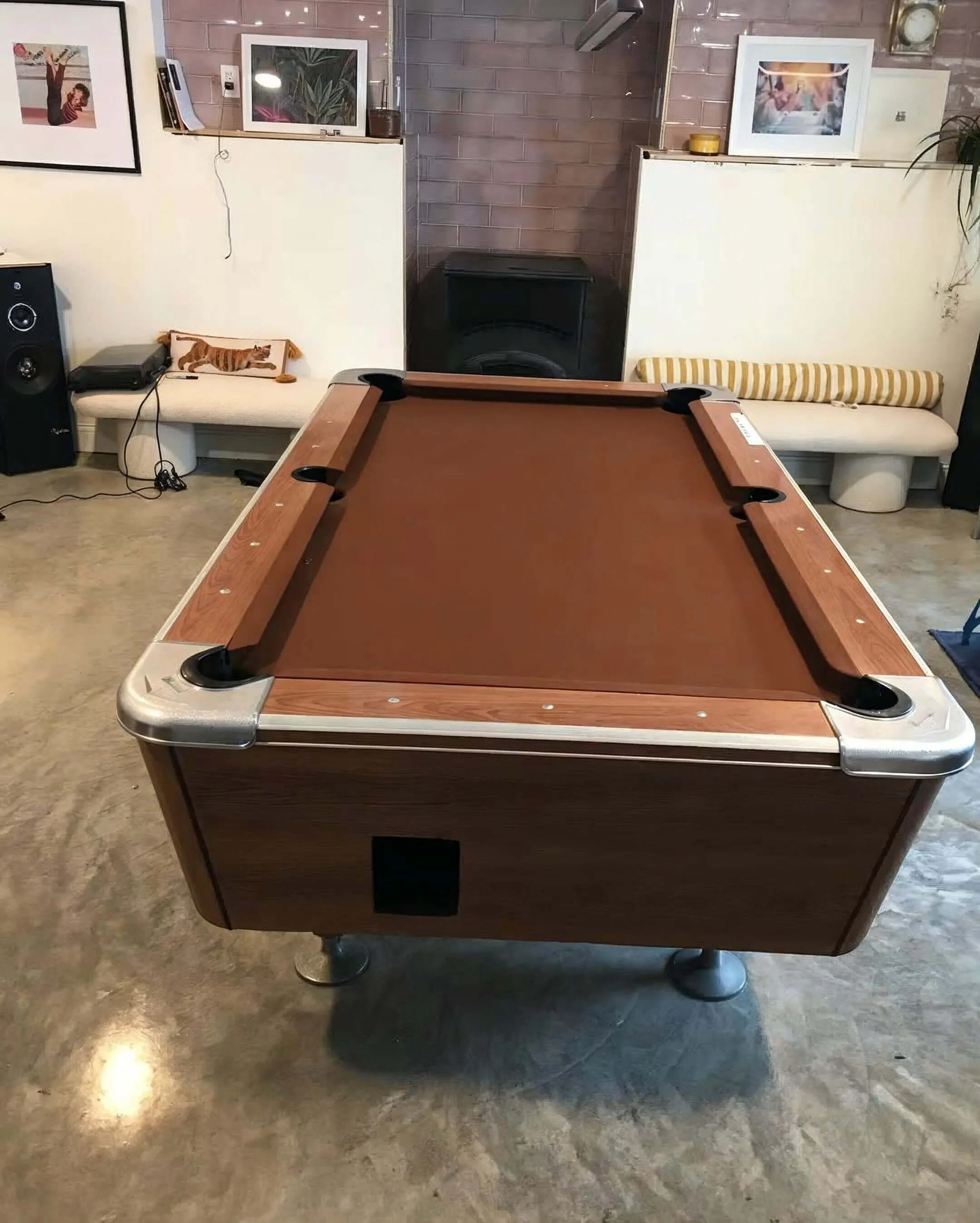 A pool table with a brown felt surface and wooden sides is in the center of a room with a concrete floor. The room has artwork hanging on the walls, two white benches with cushions on either side of a black speaker, and various decorative items. Ther