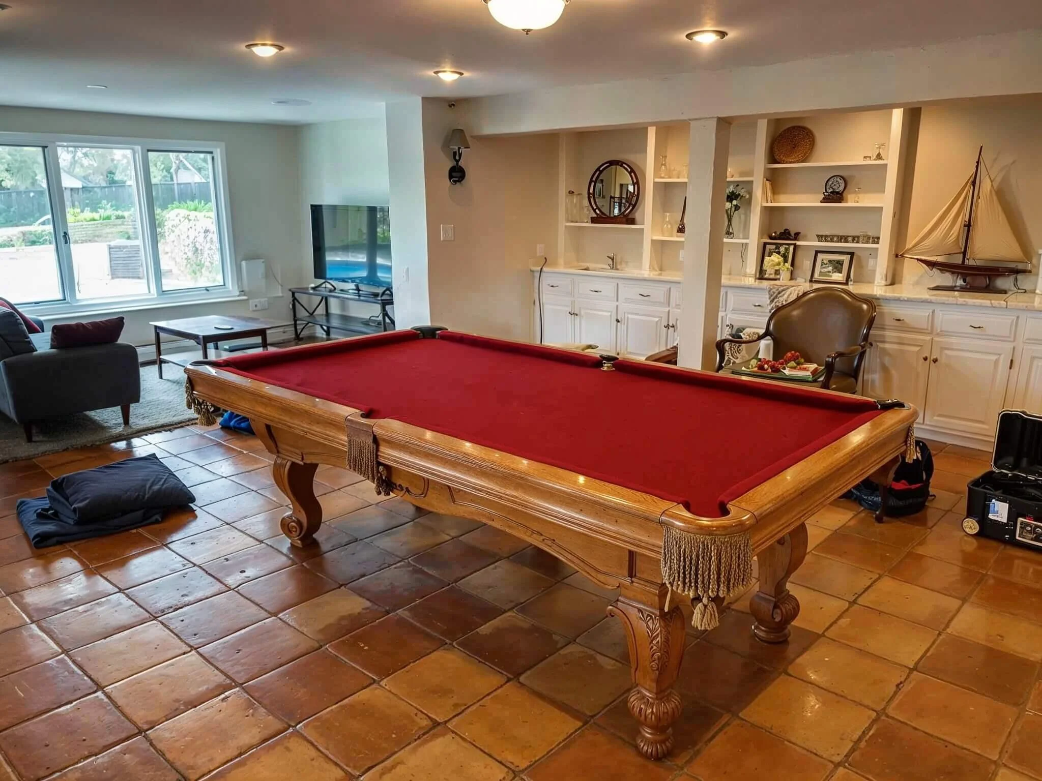 A room with a red felt pool table on a tiled floor, a sitting area with dark couches by a window, and a built-in white bookshelf and cabinets behind the pool table with decorative items. There is a leather armchair and a model sailboat on the right s