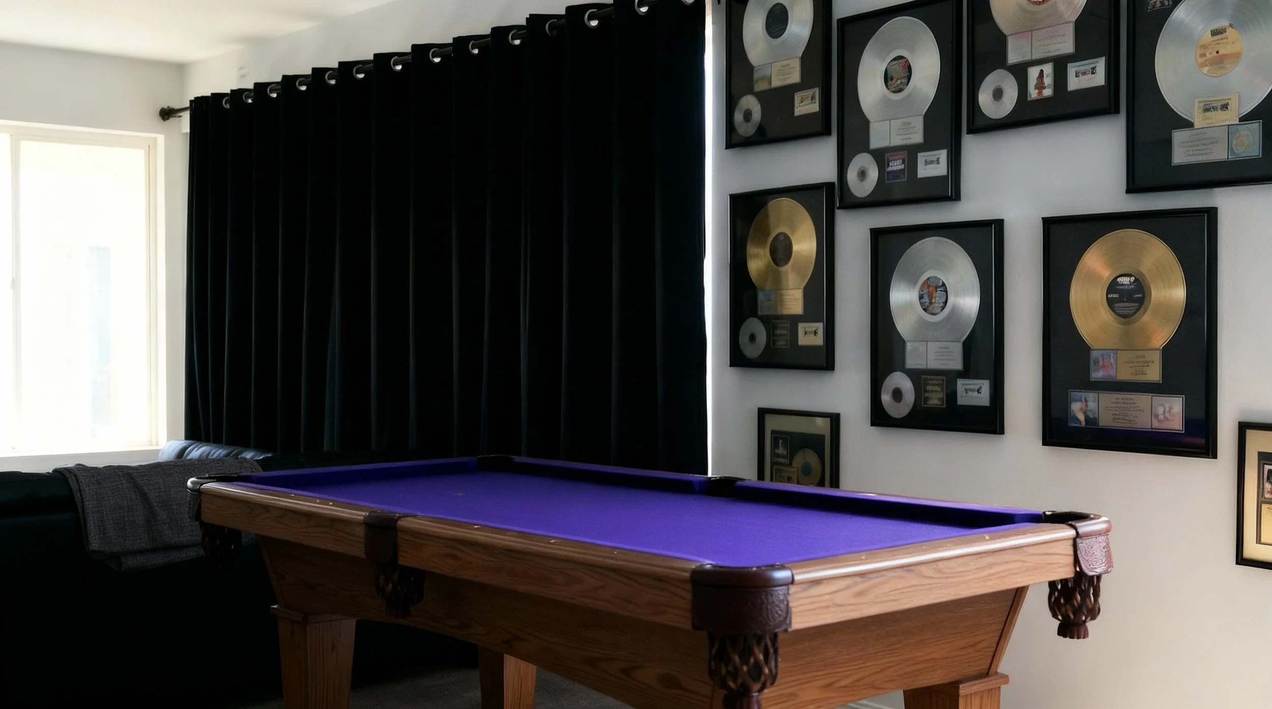 A room with black curtains on the left side, a window with natural light, a leather sofa with a blanket on it, and a wooden pool table with a purple felt surface in the center. The wall on the right side displays multiple framed awards and gold and s
