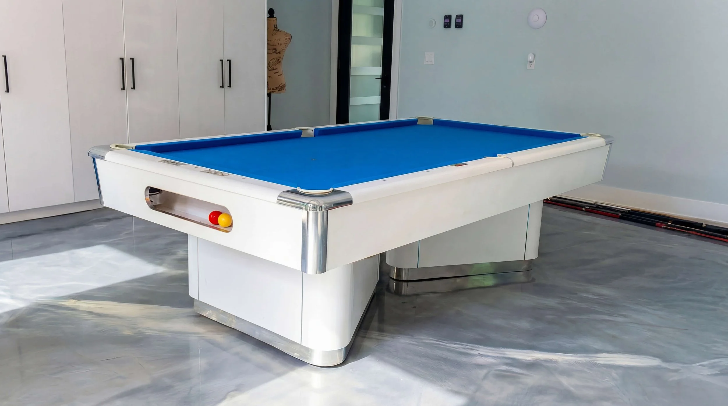 Indoor air hockey table with blue playing surface and red and yellow mallets on the side, in a modern room with white cabinets and gray flooring.