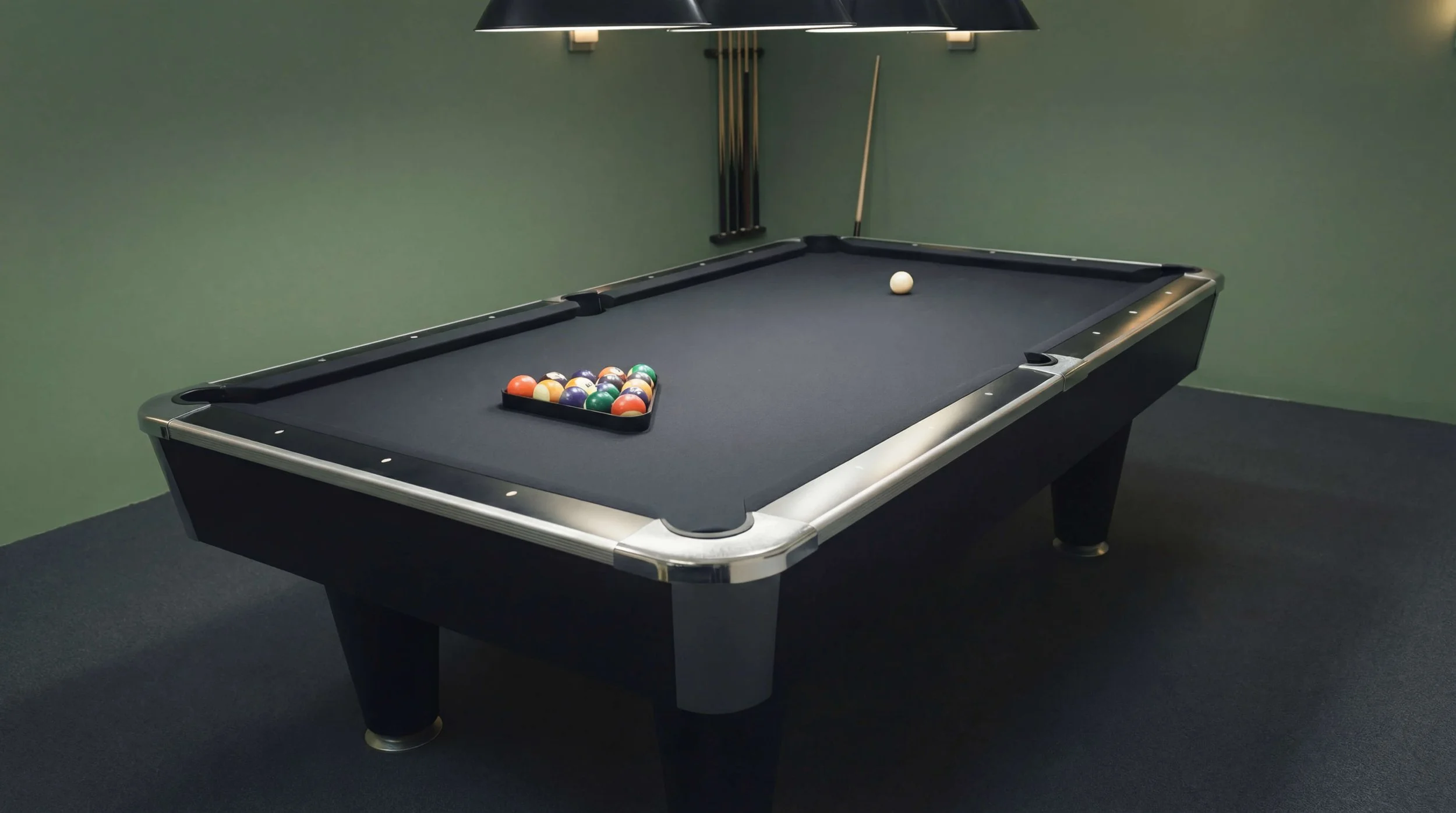 Black pool table with balls racked, cue ball, and cue sticks hanging on the wall in a room with green walls and black carpet.