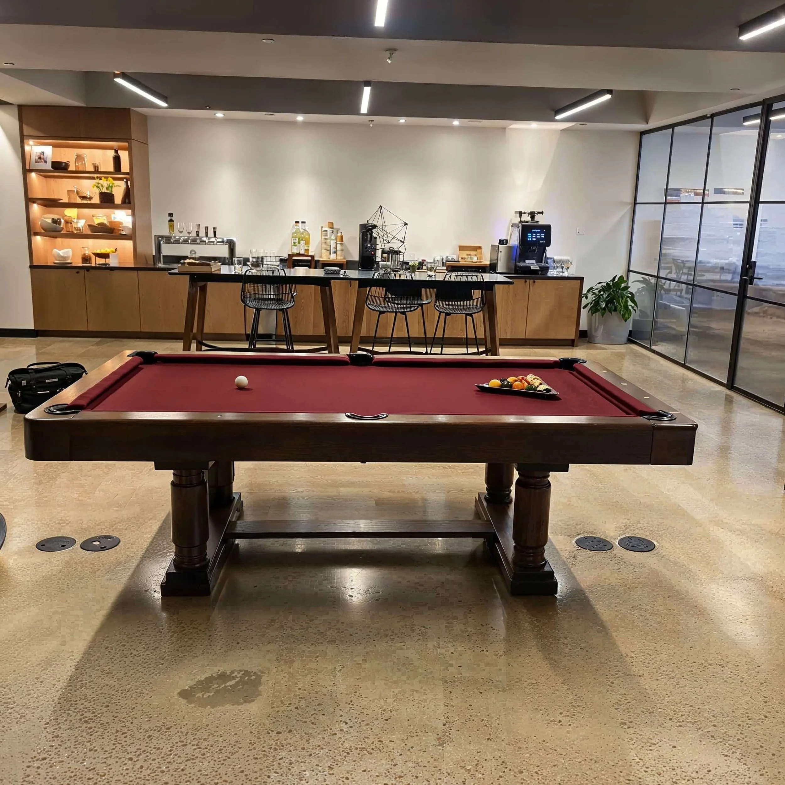 Indoor lounge with a red-felt pool table set up for a game, with a ball and a rack of balls on the table. In the background, there is a bar area with bar stools, a coffee machine, and shelves with bottles and glasses. Large windows with a view outsid
