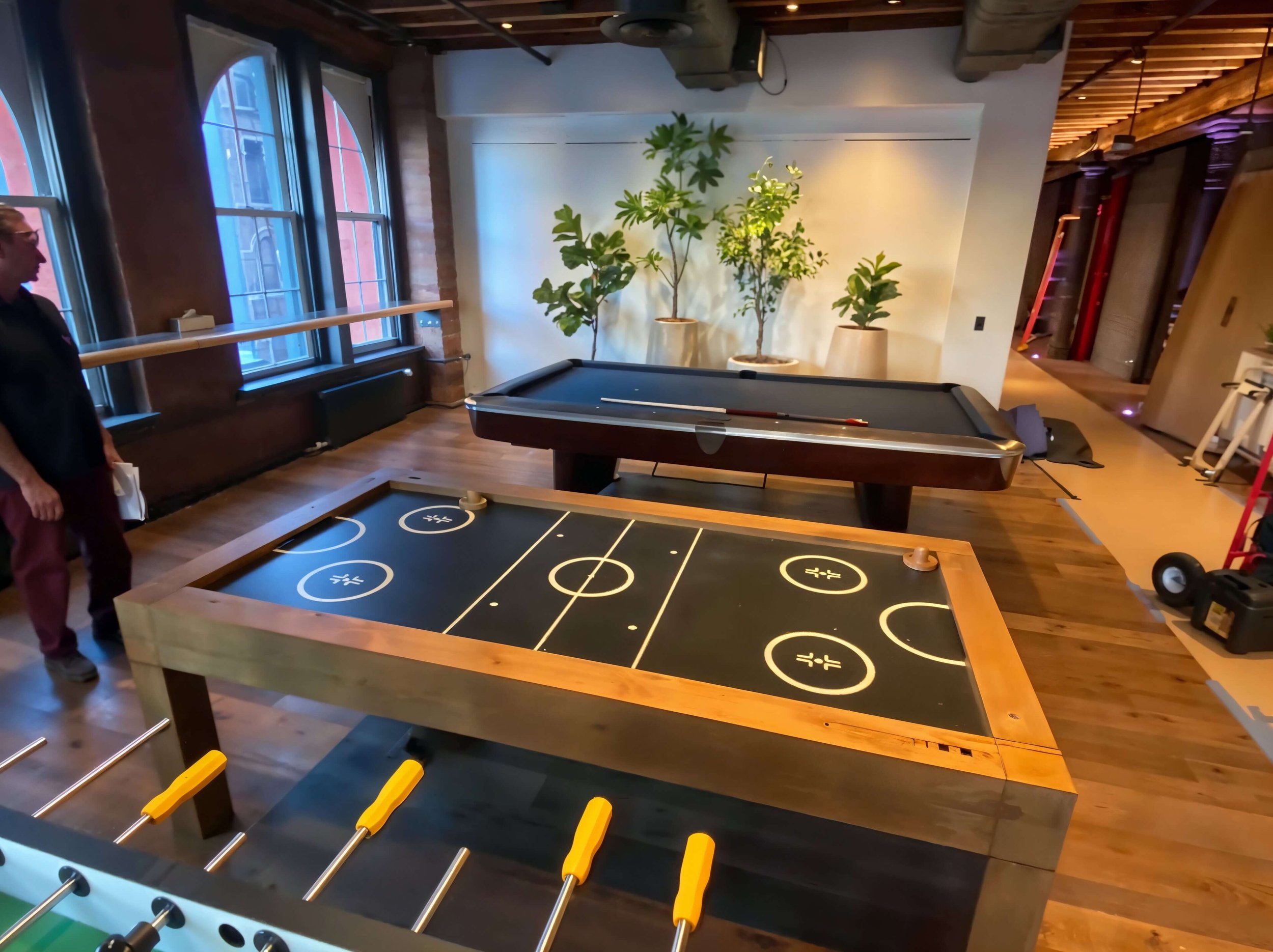 Indoor recreational room with a shuffleboard table in the foreground, a foosball table, and an air hockey table in the background. Large windows with a view of brick buildings outside, wooden floors, and a wall decorated with potted plants.