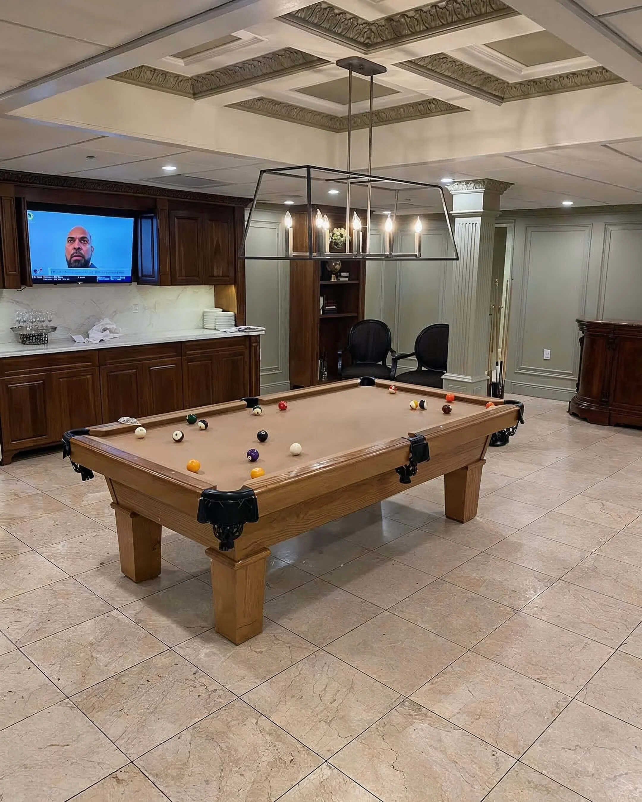 Interior of a room with a pool table, a wall-mounted TV playing a man's face, and a chandelier hanging above the pool table.