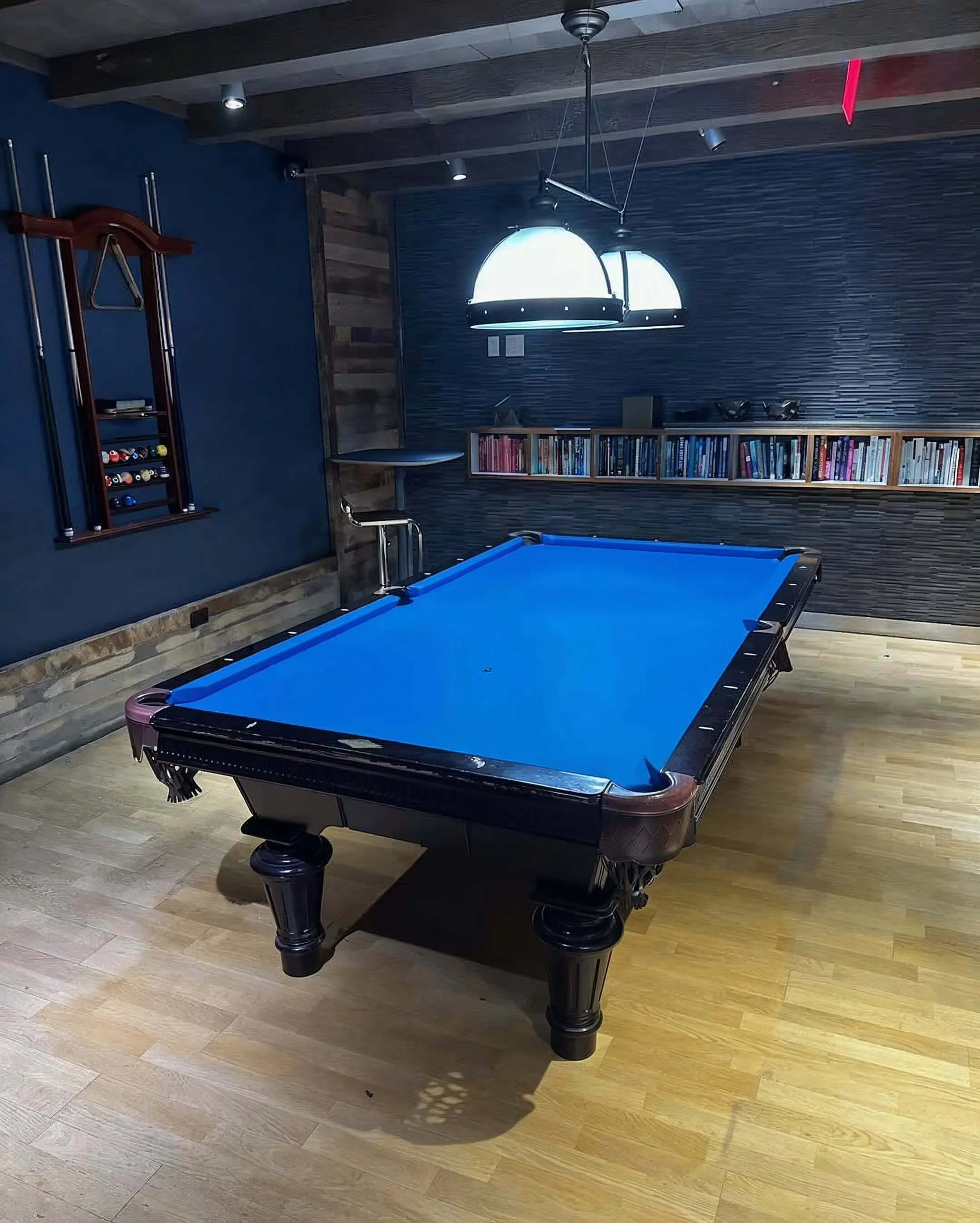 A room with a blue pool table, a wooden bookshelf with books mounted on the wall, modern ceiling lights, and a dark wood panel wall with a textured finish. The floor is light wood, and there is a small table and a rack for cues on the left wall.