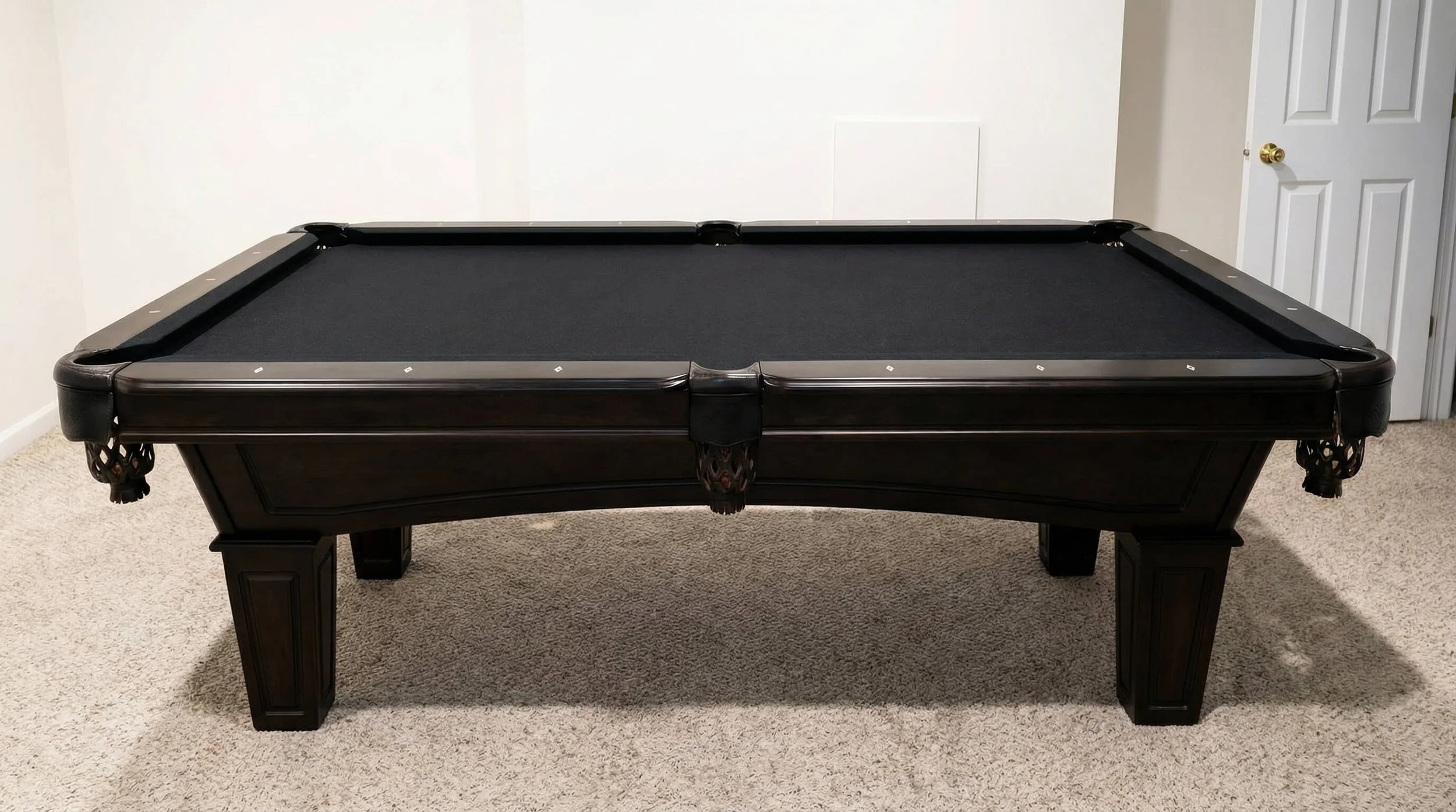 Black pool table with a black felt surface in a room with beige carpet and white walls, open white door in the background.