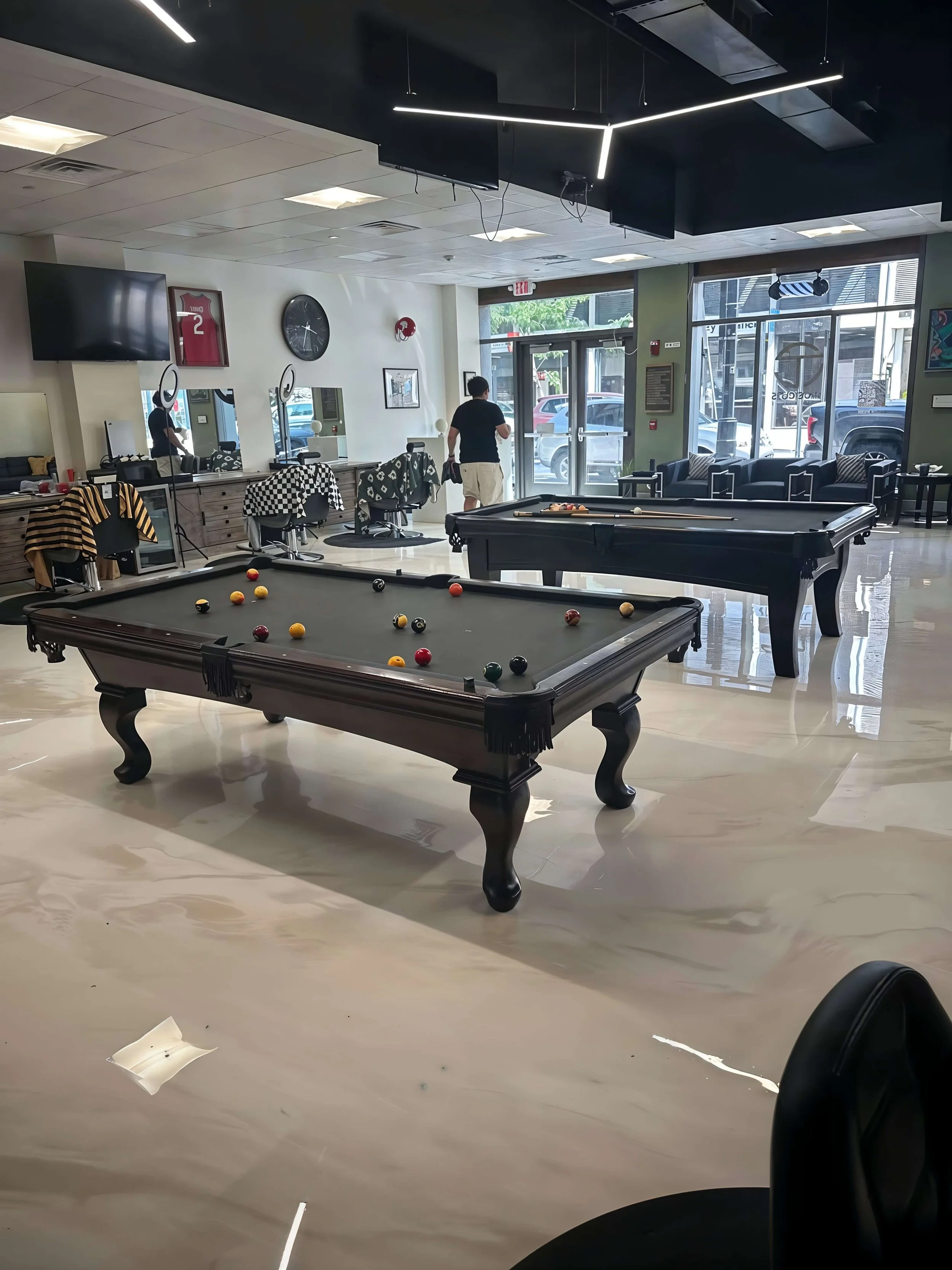An indoor lounge with two pool tables, a few people, and large windows showing parked cars outside.