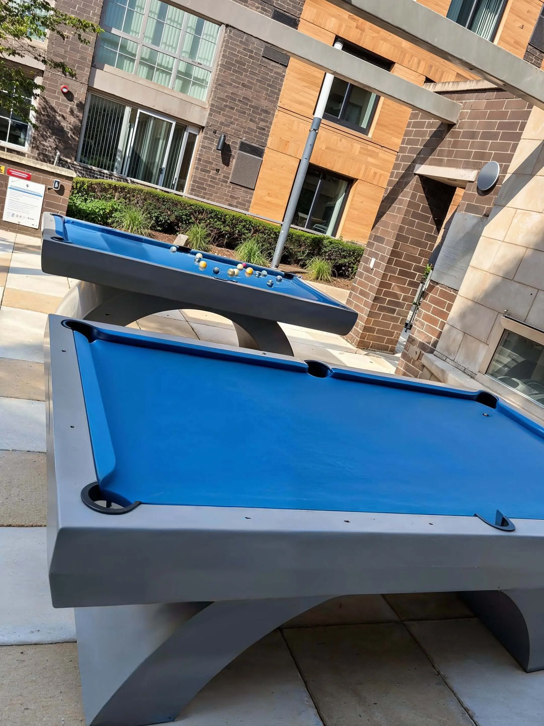 A pool table and a foosball table in an outdoor area of an apartment complex. The pool table is blue with cue slots, and the foosball table is also blue with billiard balls on it, set against a backdrop of modern apartment buildings with brick and wo