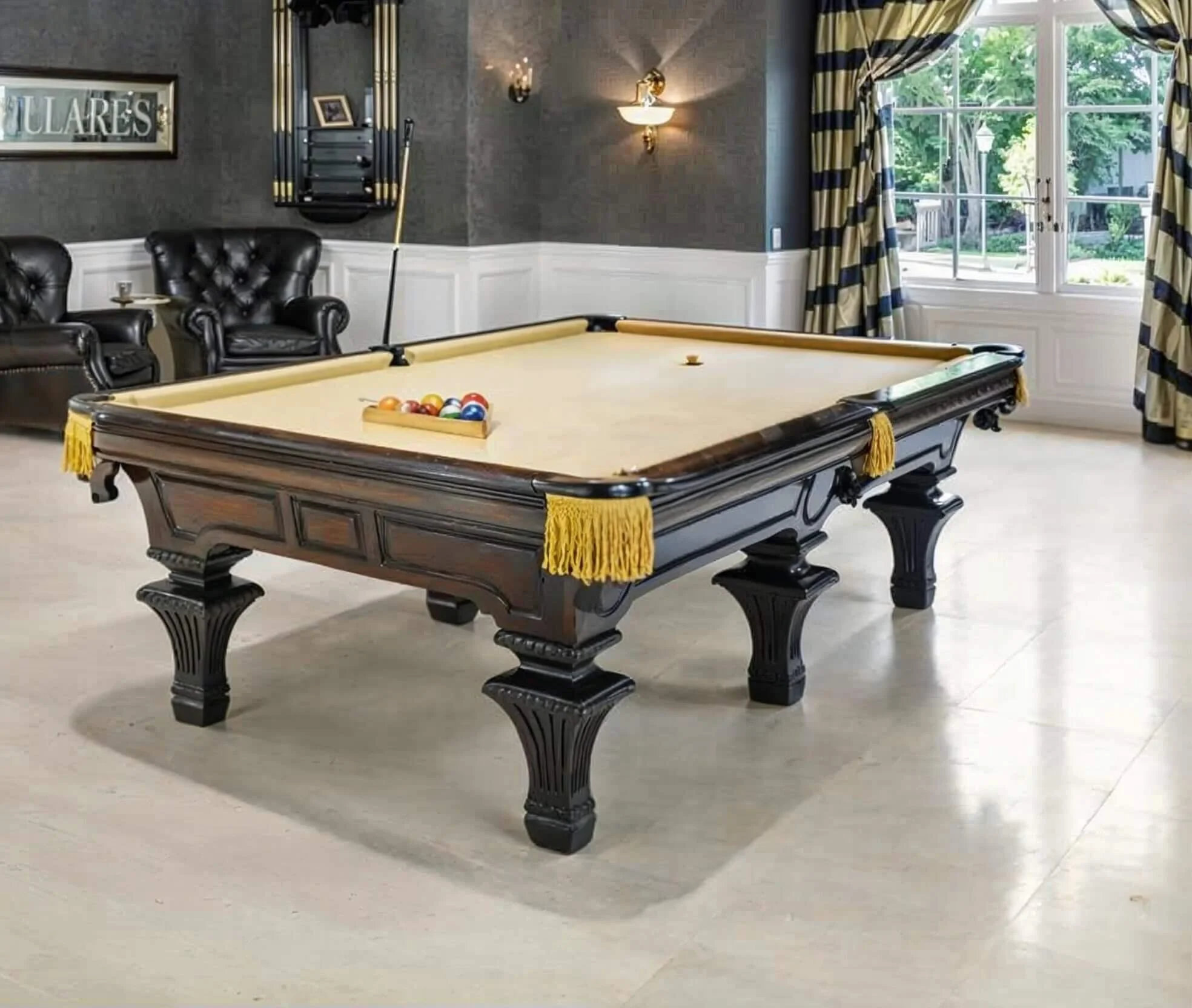 Pool table in a well-lit room with black leather chairs, a window with blue and yellow curtains, and walls decorated with framed pictures and a wall-mounted shelf.