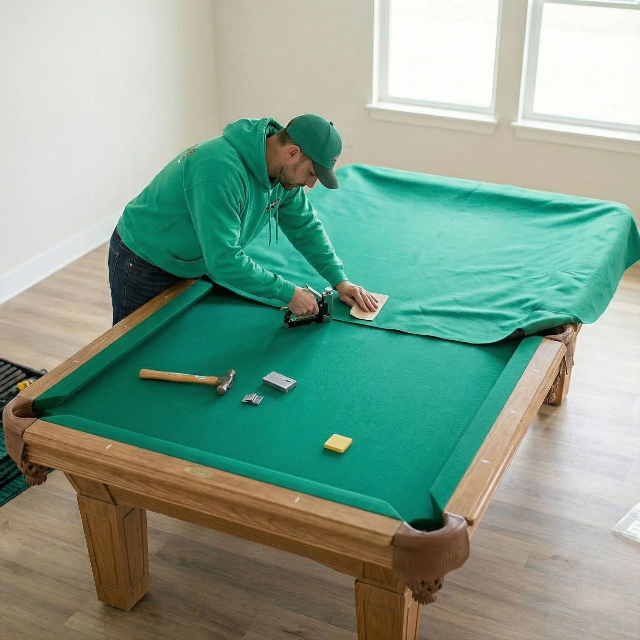 A man wearing a green hoodie and cap is upholstering a pool table with green fabric, using a staple gun, in a well-lit room with wooden flooring and windows.