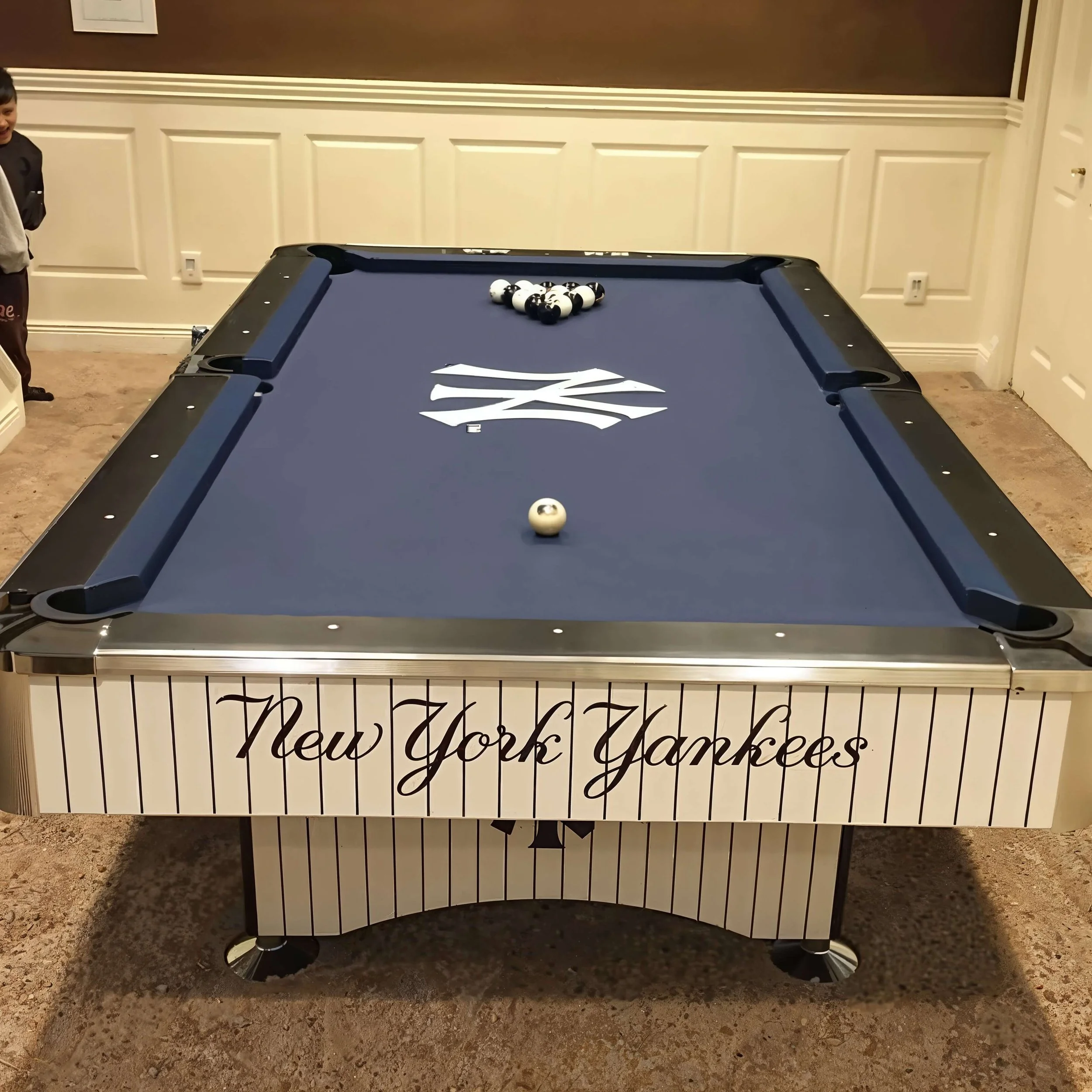 A pool table with a New York Yankees logo on the felt, pool balls racked at the top, and a single ball in the middle of the table. A boy is partially visible on the left side of the image.