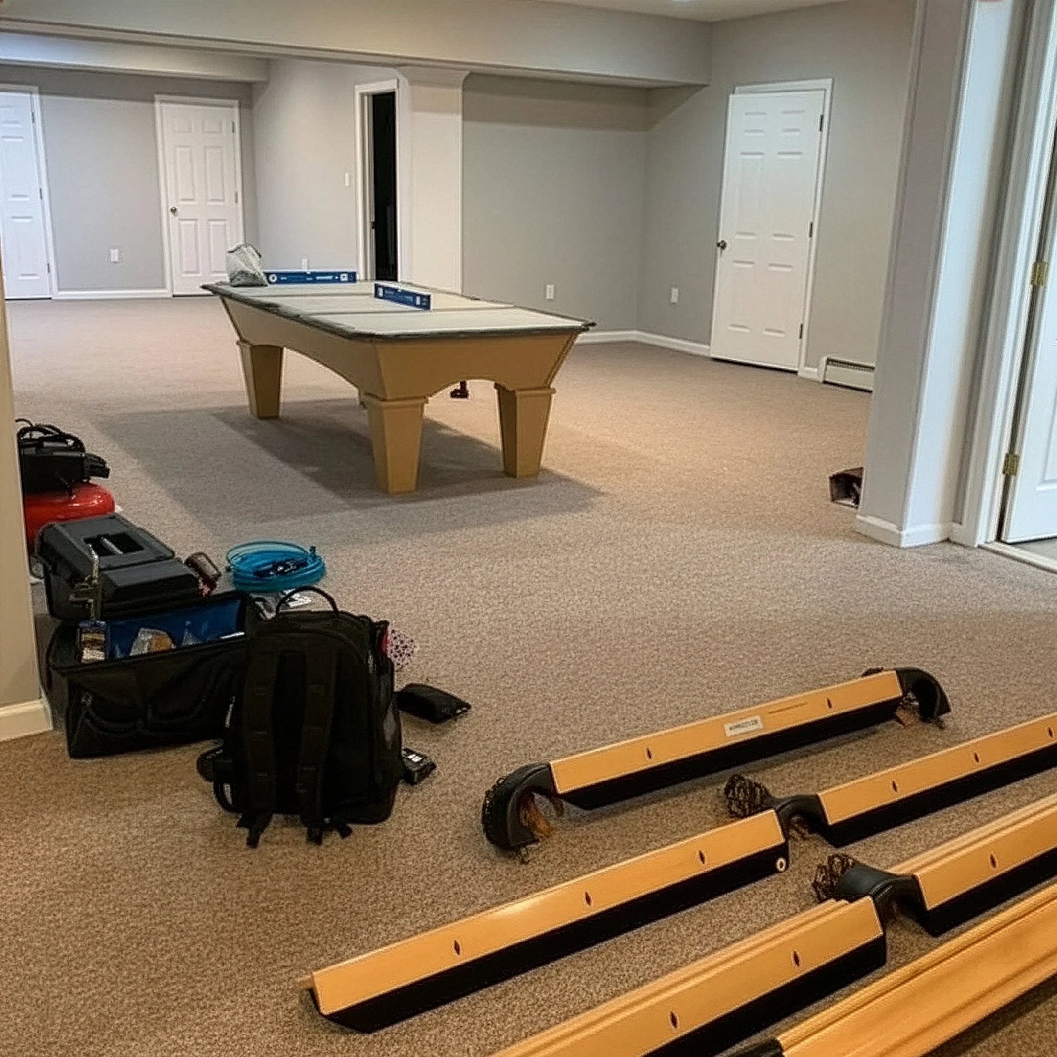 Empty room with beige carpet, folding table, and some tools and equipment on the floor, including desk frames, bags, and boxes, suggesting ongoing setup or renovation.