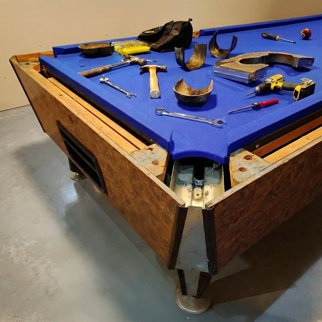 Disassembled vintage pool table with blue felt top, tools, and parts scattered on it, in a workshop or garage setting.