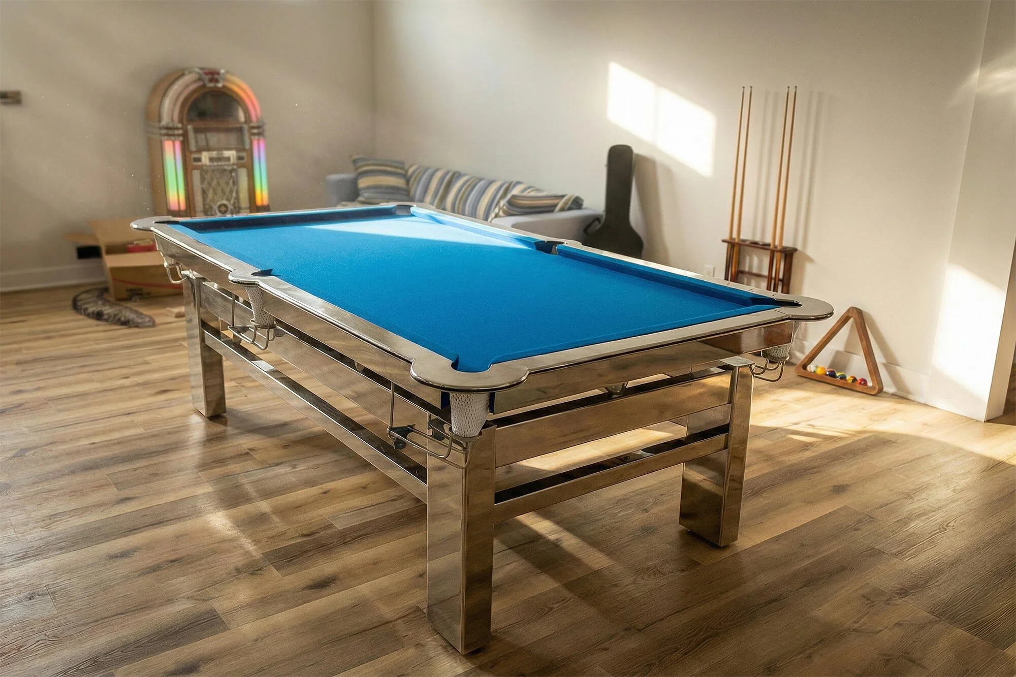 A metal pool table with a blue felt surface in a room with wooden flooring, a striped couch, a guitar case, a vintage jukebox, and a triangle-shaped rack with billiard balls.