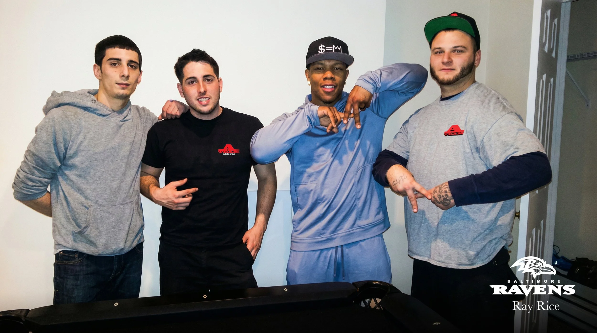 Four men standing together, one wearing a gray hoodie, one in a black t-shirt, one in a gray and black baseball cap and gray tracksuit, and one in a gray Ravens shirt with tattooed arms, posing for a photo indoors with the Baltimore Ravens logo and t