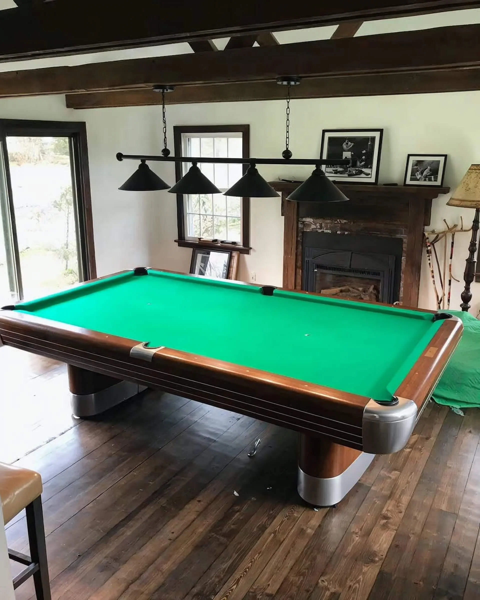 A room with a green pool table, a fireplace, and black hanging lights above the table. There are windows and framed photographs on the wall.