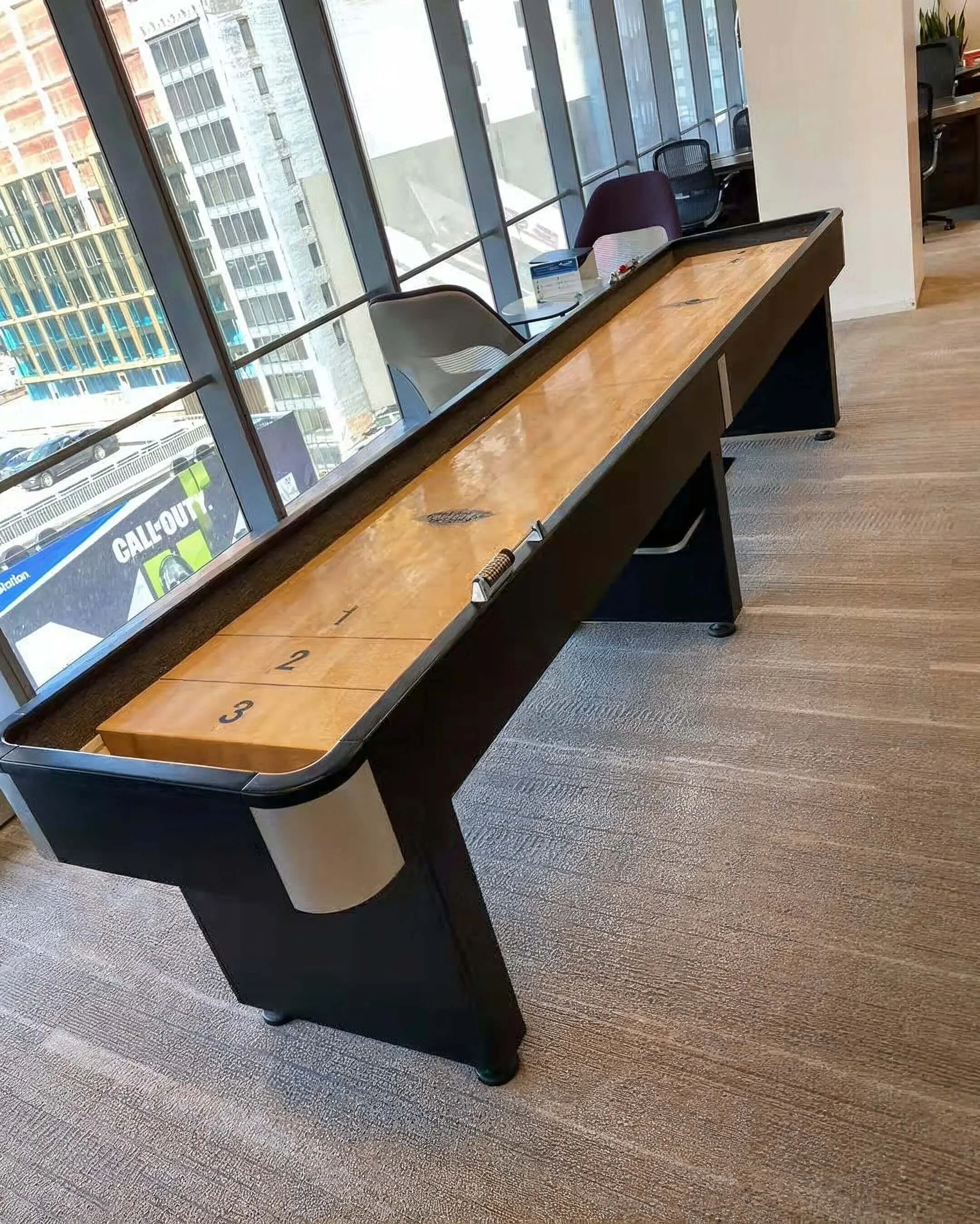 Shuffleboard table in an office, numbered sections 1, 2, and 3, located near a large window with city buildings outside.