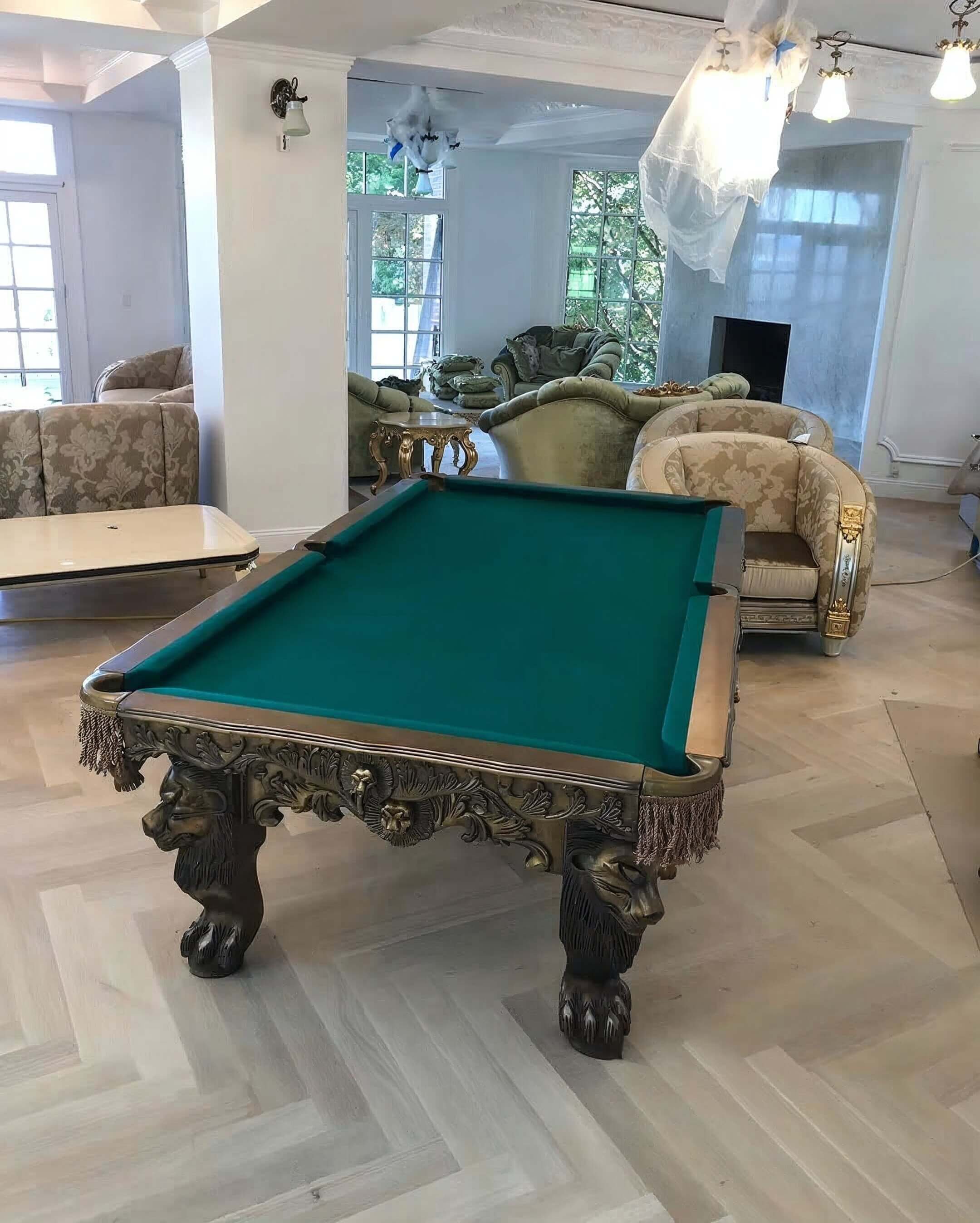 A living room with an ornate, carved pool table with green felt and lion paw legs, surrounded by vintage-style upholstered armchairs and sofas. There are large windows, a fireplace, and some ceiling lights.