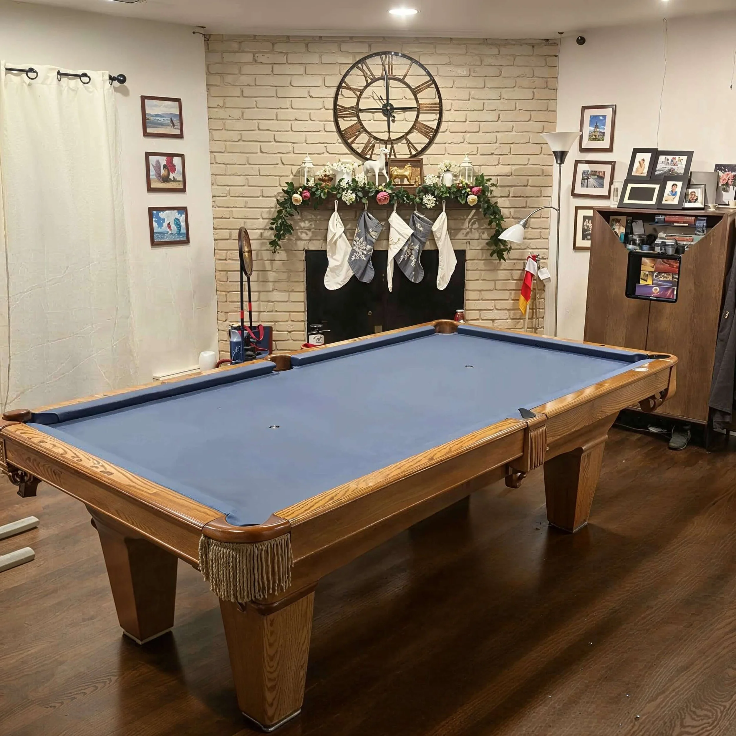 A cozy room with a blue pool table, hanging stockings on a fireplace mantel, and a large clock above the mantel decorated with flowers and small ornaments, surrounded by family photos on the walls.