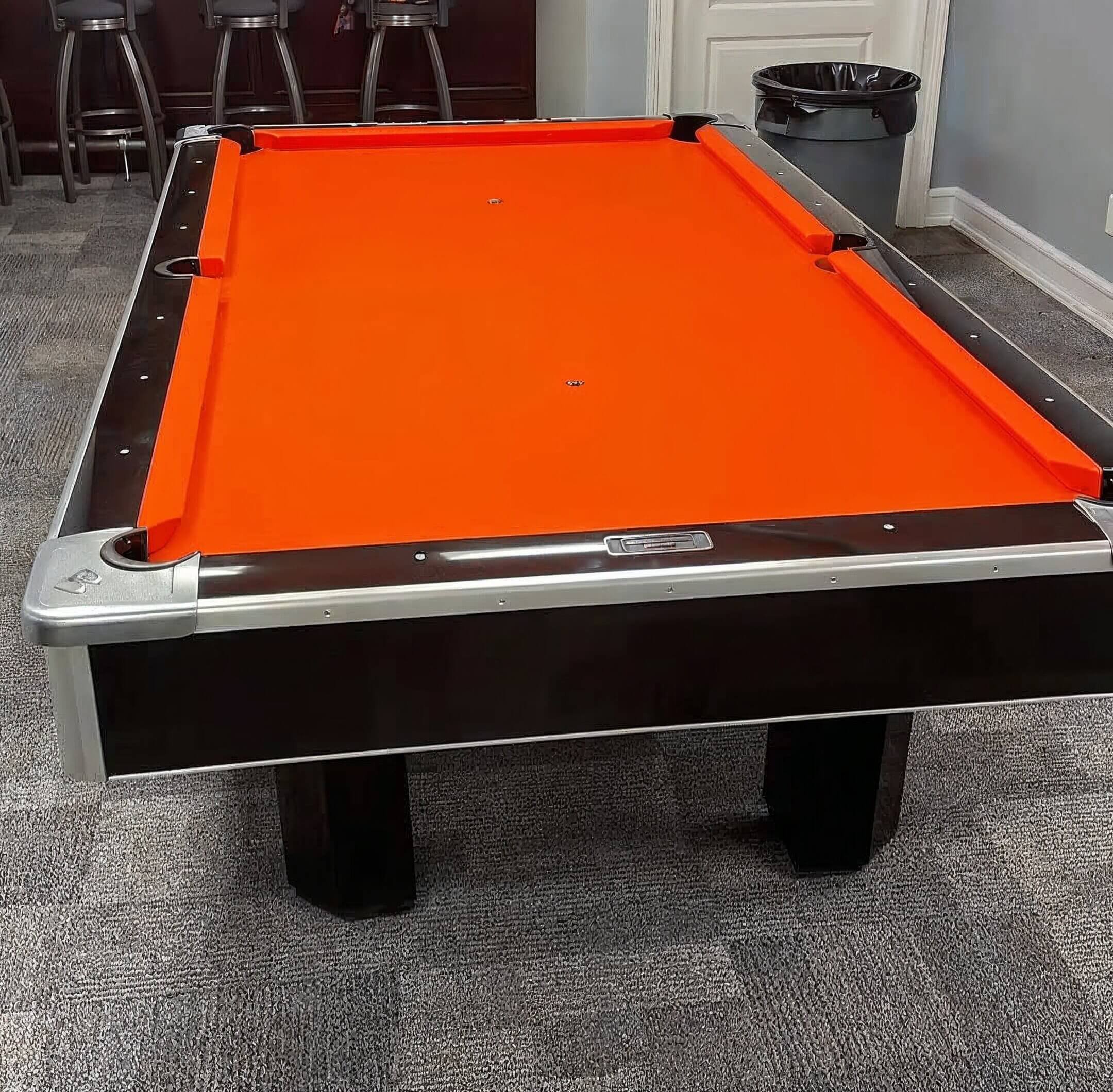 An indoor billiards table with a bright orange felt surface, black sides, and corner pockets, situated on a gray carpeted floor in a room with some bar stools and a trash can in the background.