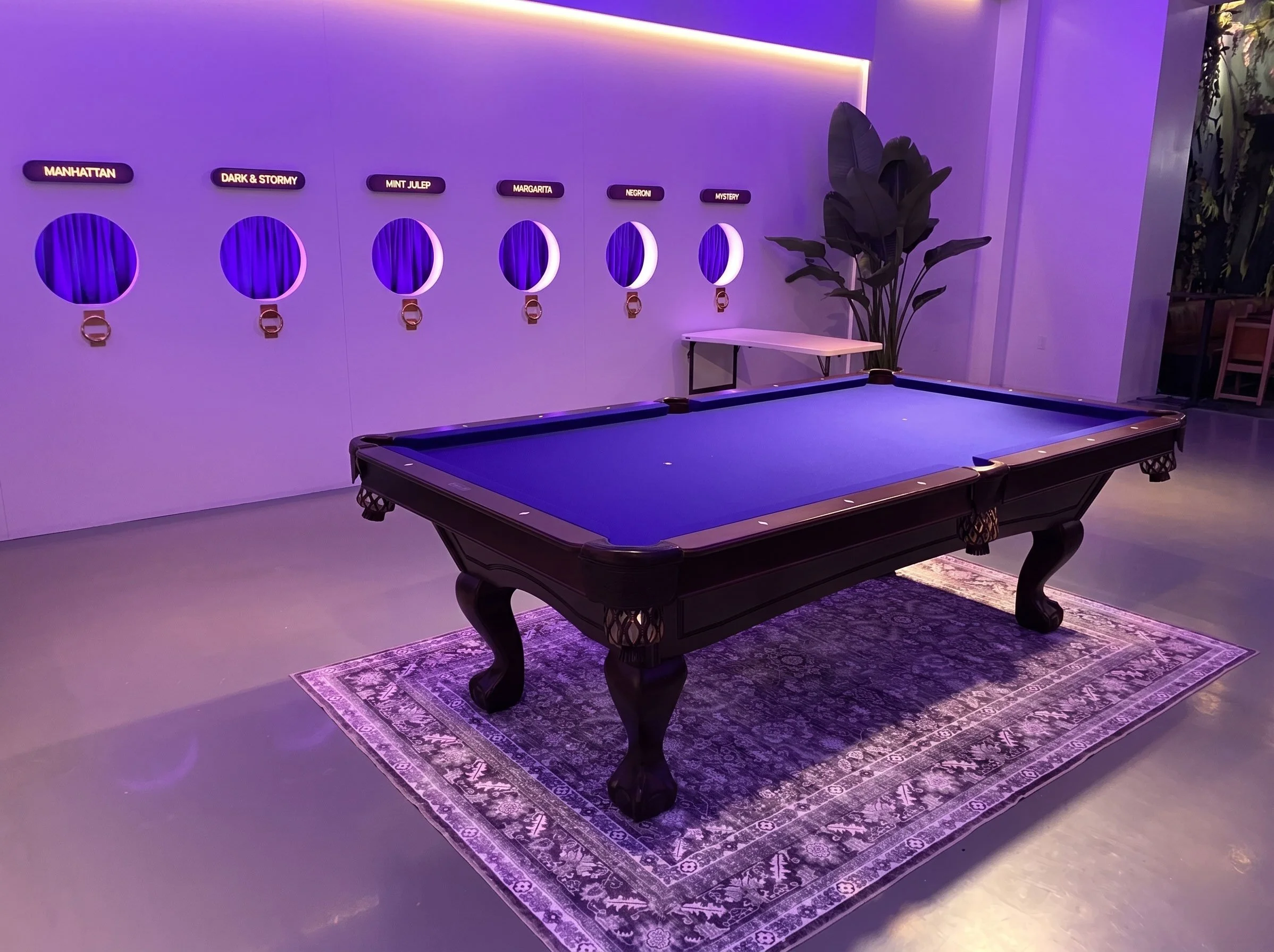 Indoor billiards room with purple lighting, a pool table on a patterned rug, wall-mounted illuminated signs with names of cocktails, potted plant, and purple drapery.