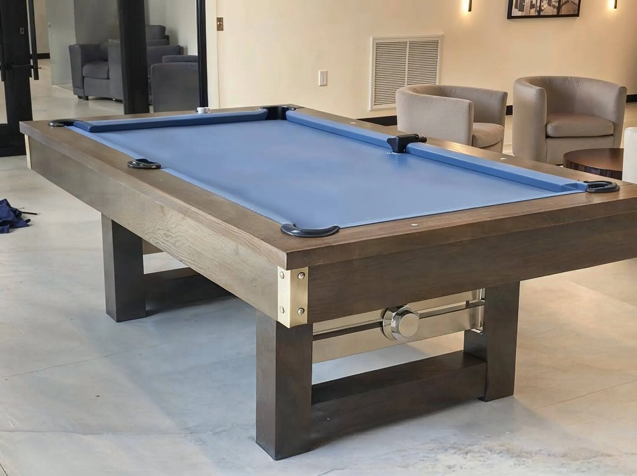 A pool table with a blue playing surface and wooden frame in a lounge area with beige chairs and a small round table.
