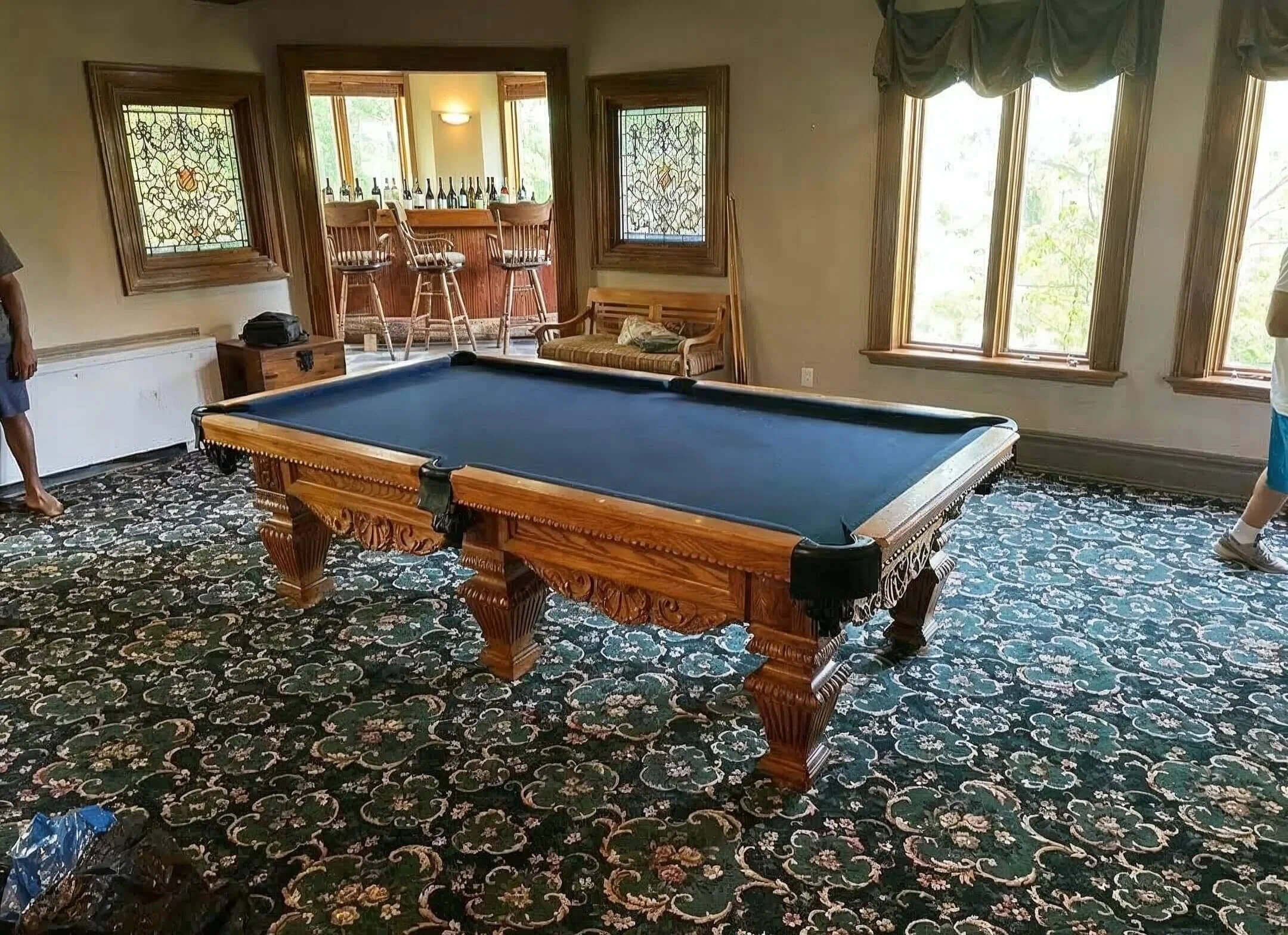 A room with a pool table featuring a wooden frame and blue felt, surrounded by windows with wooden trim, dark floral carpet, and a bar area with stools in the background.